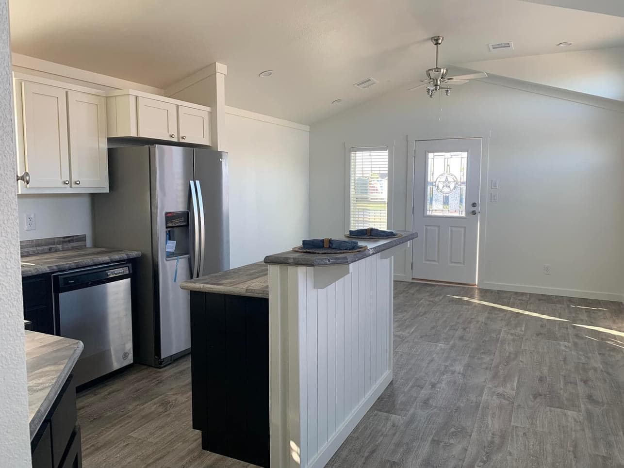 Bright kitchen with white walls, vaulted ceiling, and large window. Features stainless steel appliances, gray countertops, and wood-like flooring. Cozy and modern ambiance.