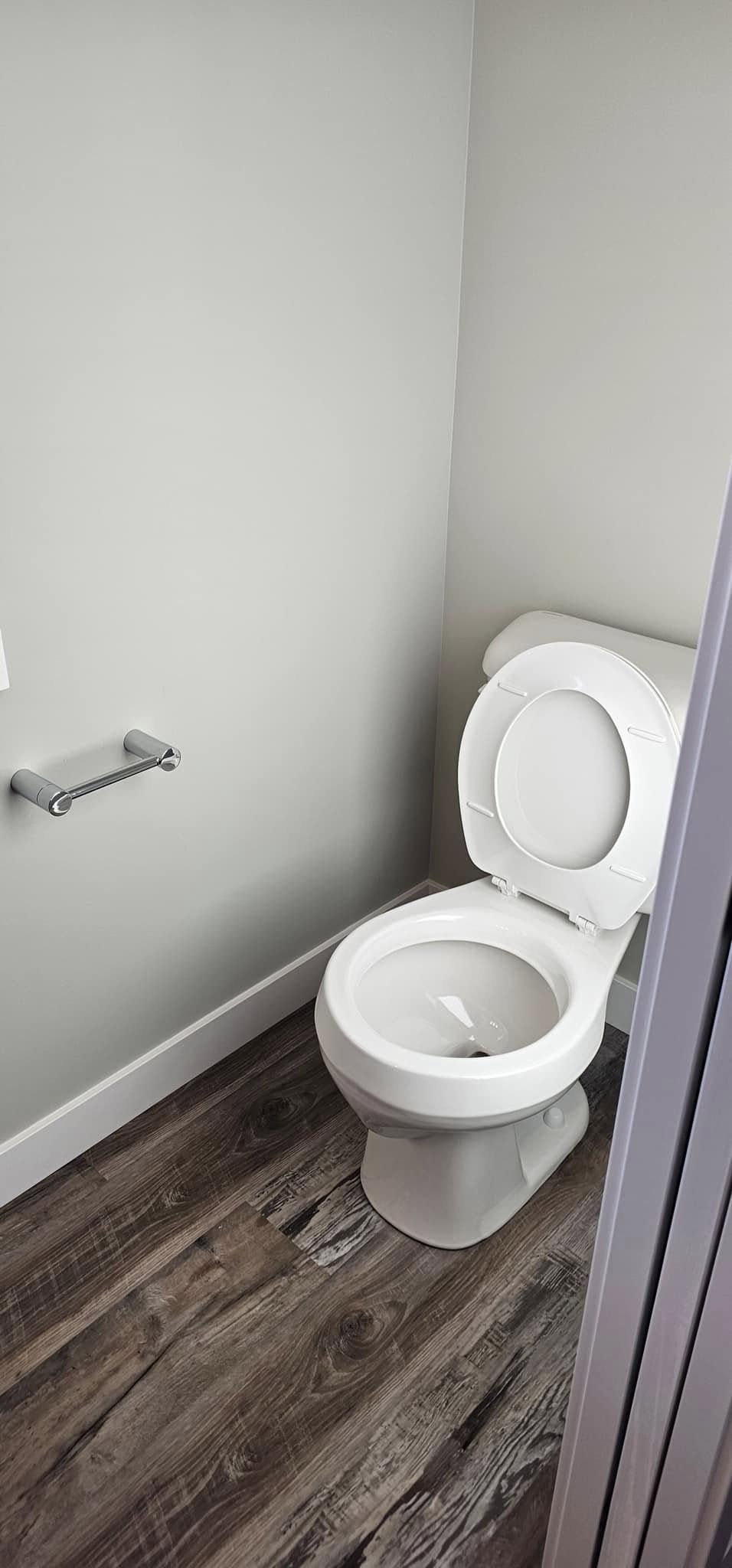A clean, white toilet with an open lid is positioned against a light gray wall. To the left is a silver toilet paper holder without a roll. The floor is dark wood, creating a simple, modern, and tidy bathroom scene.