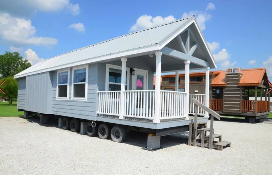 A small, light blue tiny house on wheels with a white porch sits on a gravel lot under a clear blue sky. Other small houses are visible in the background.