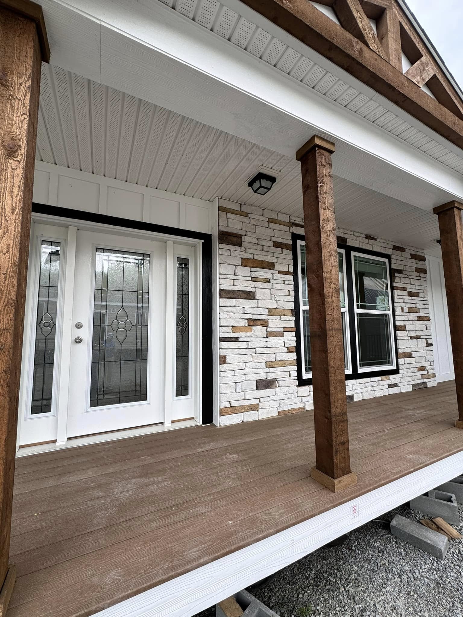 Front porch of a house with brown wooden columns and a white textured ceiling. The facade includes a white brick wall and two large windows.