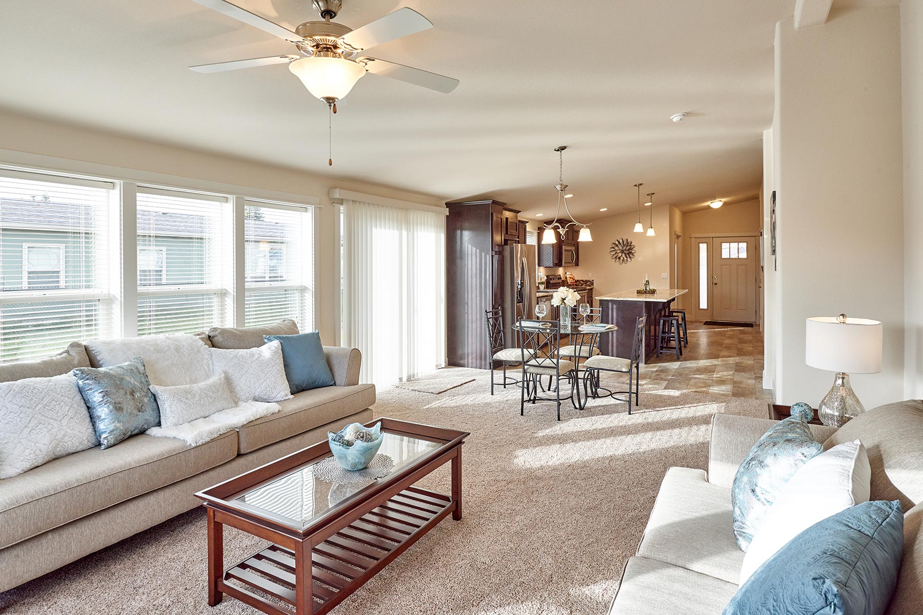 Bright, open-plan living room and kitchen with beige sofas, glass coffee table, ceiling fan, and dining area. Sunlight filters through large windows. Cozy and inviting tone.