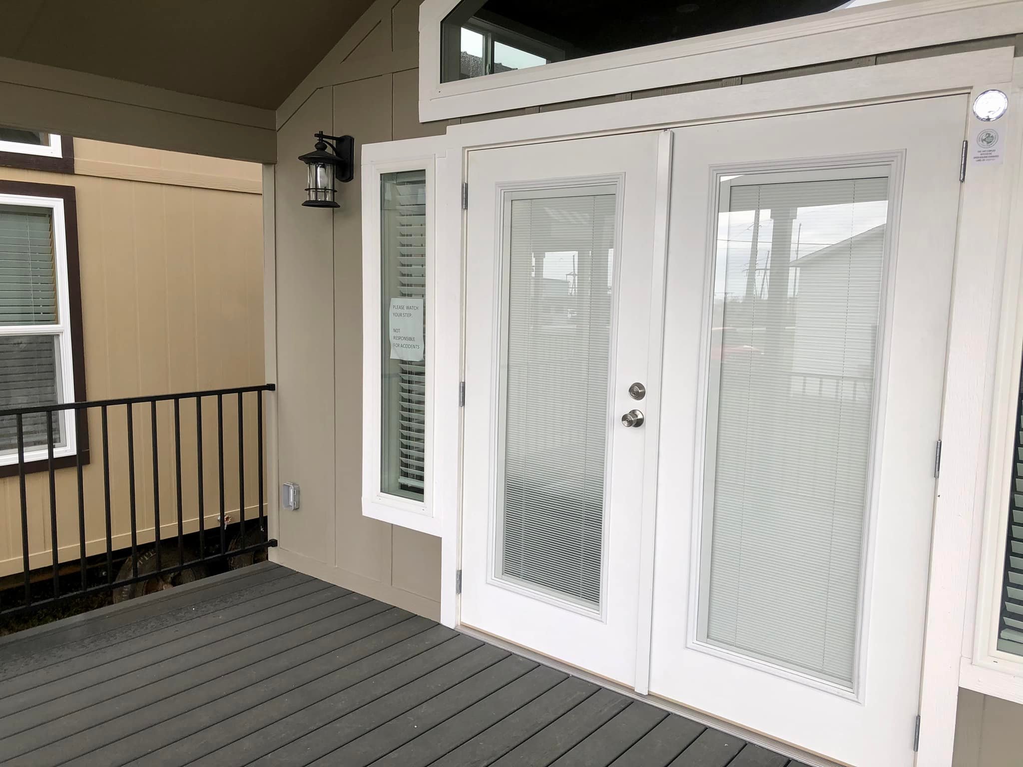 A beige house exterior featuring double glass doors with white frames and blinds. A black wall lamp is mounted beside the door. A black metal railing is visible on the left.
