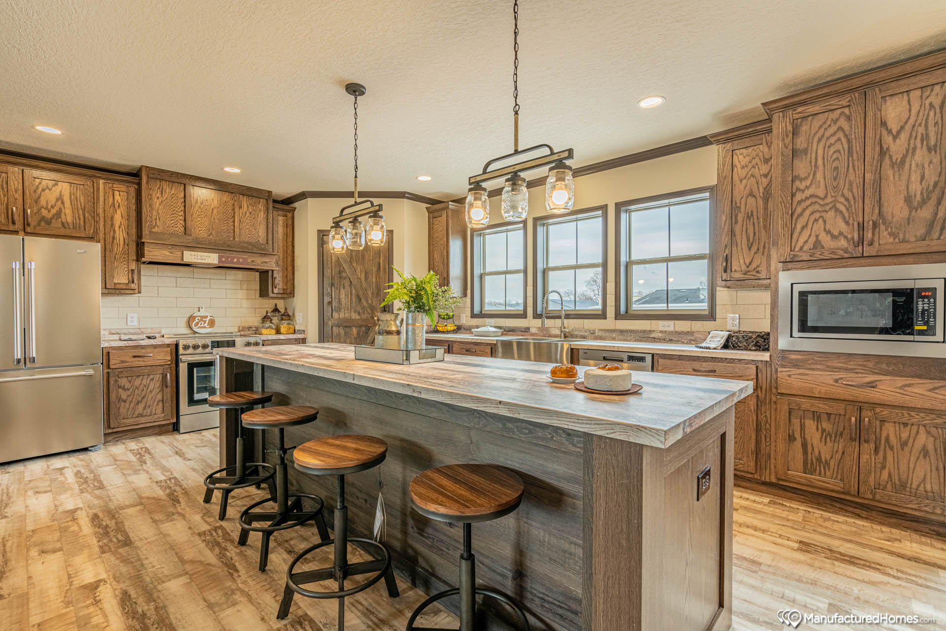 Spacious kitchen with rustic wood cabinets, large island with wooden stools, stainless steel appliances. Pendant lights provide warm ambiance.