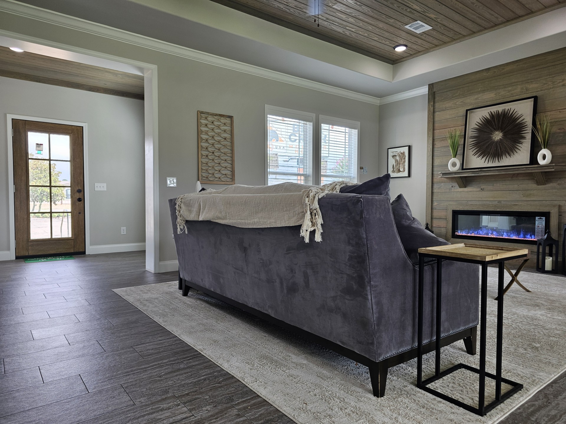 Modern living room with a gray couch and beige throw, wooden accent wall, electric fireplace, and artwork. Bright, welcoming tone with natural light.
