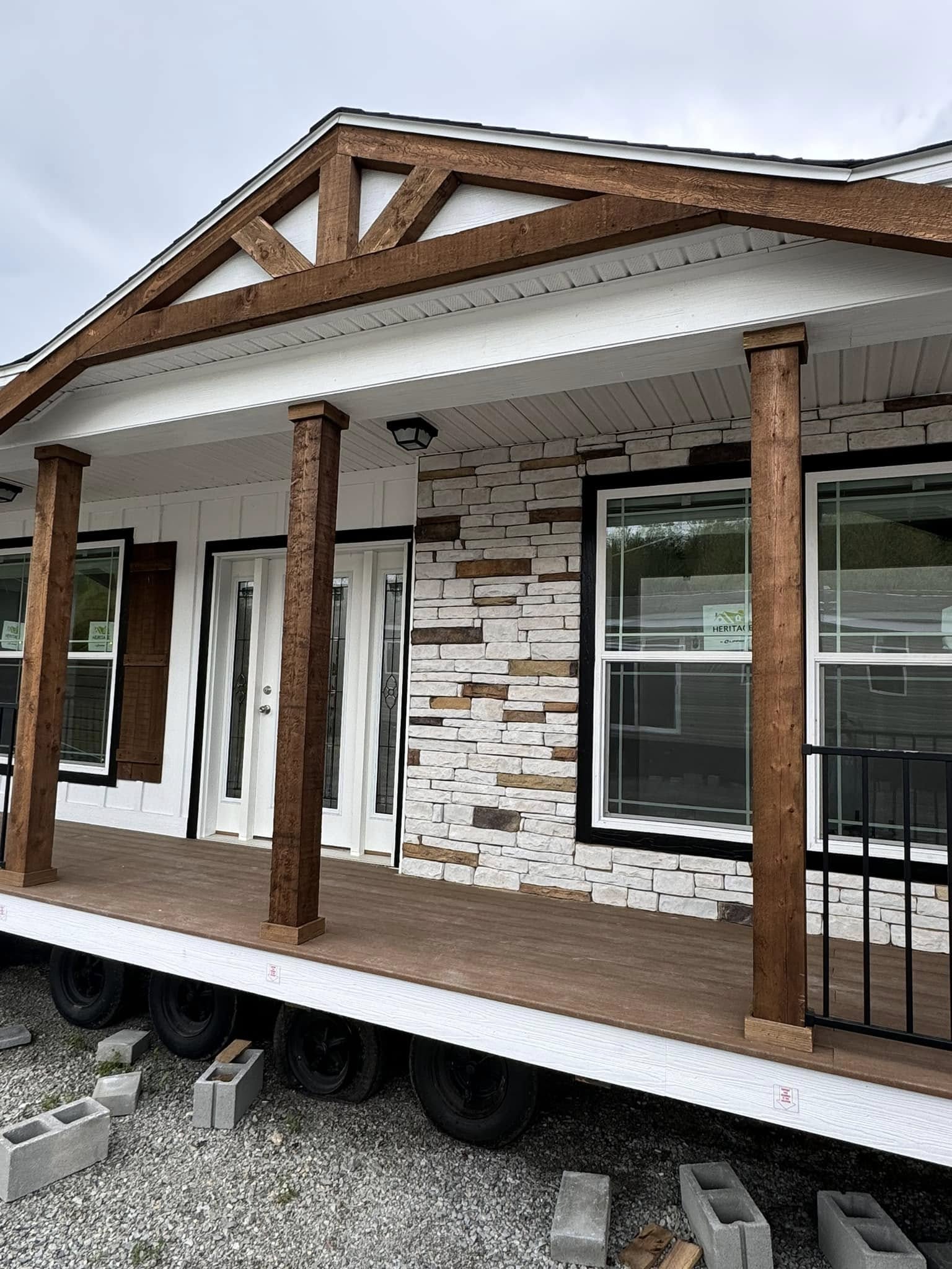 Front porch of a mobile home under construction, featuring a wooden deck, stone wall, large double doors, and a rustic wooden gable. Overcast sky.
