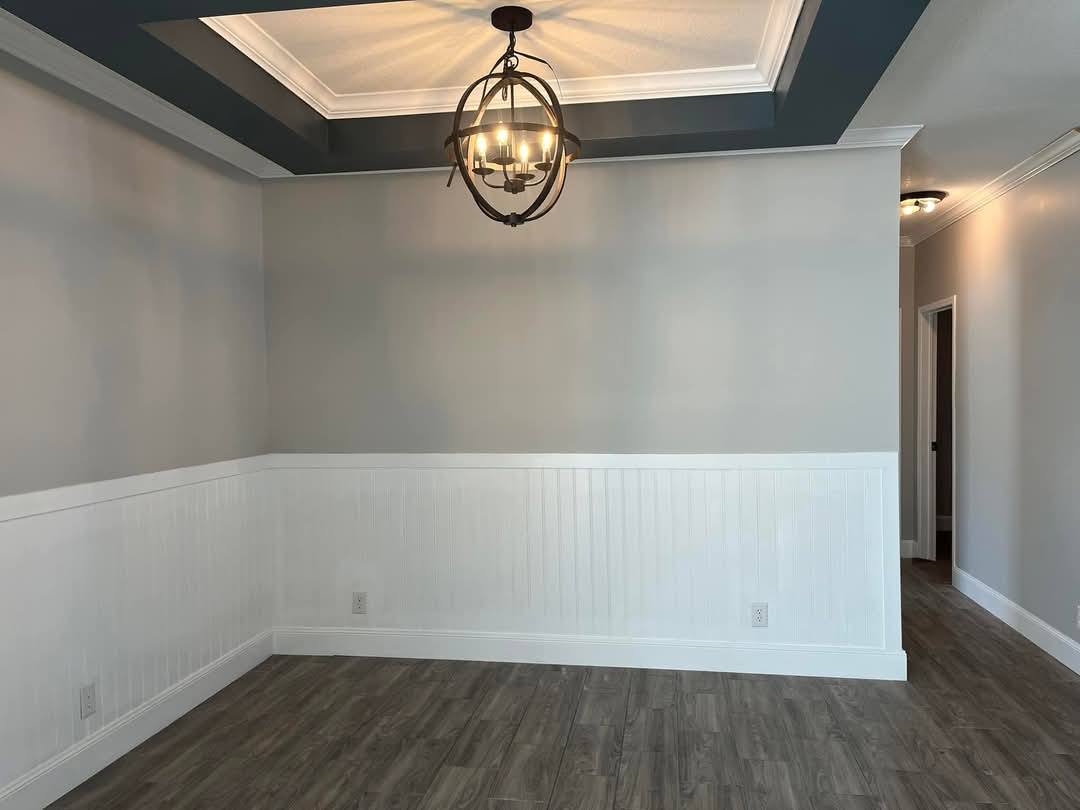 A minimalist room with light gray walls and white wainscoting, featuring a dark wood floor. A modern chandelier hangs from a recessed, coffered ceiling.