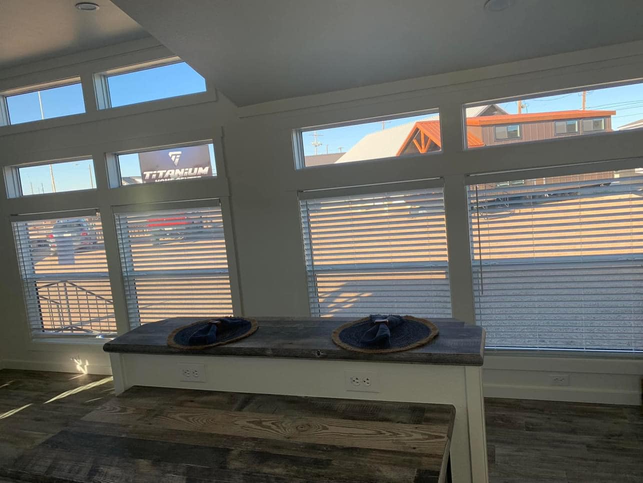 Interior view of a room with large windows covered by blinds letting in light. A wooden counter with two placemats and napkins is in the foreground.