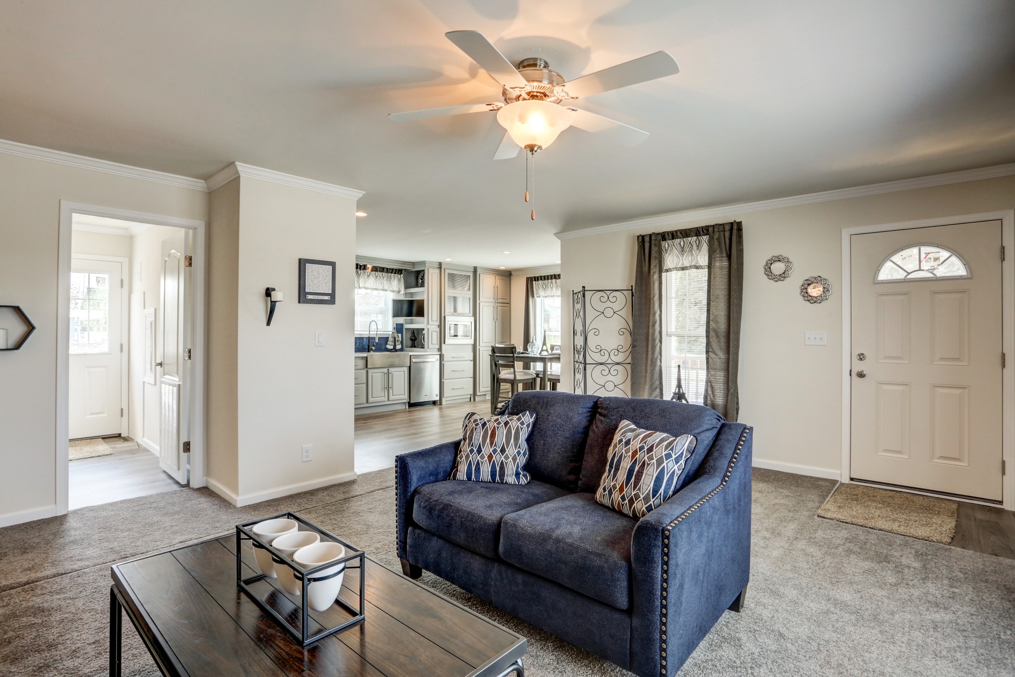 Spacious and bright living area with a navy sofa and patterned pillows. Ceiling fan above; kitchen in view. Neutral tones, cozy and welcoming ambiance.
