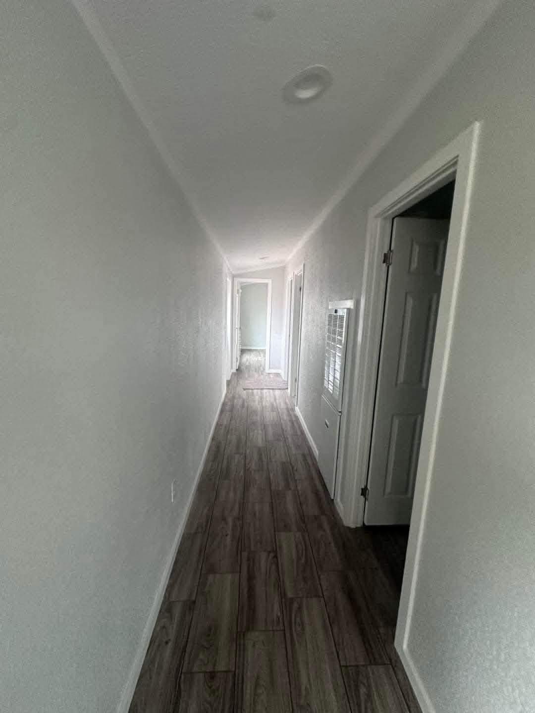 Narrow hallway with white walls and wood-patterned flooring. Several doorways open on the right. Bright light shines at the end, creating a welcoming feel.
