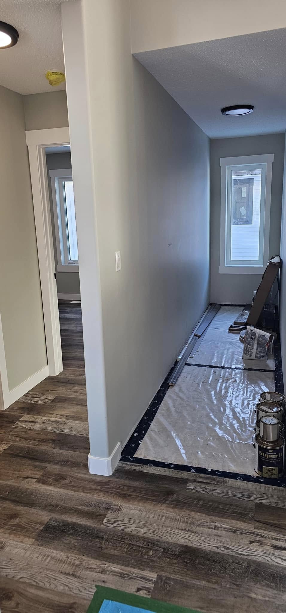 Narrow hallway under renovation with dark wood flooring, beige walls, and bright overhead lights. Windows at the end illuminate paint cans and covered floor.