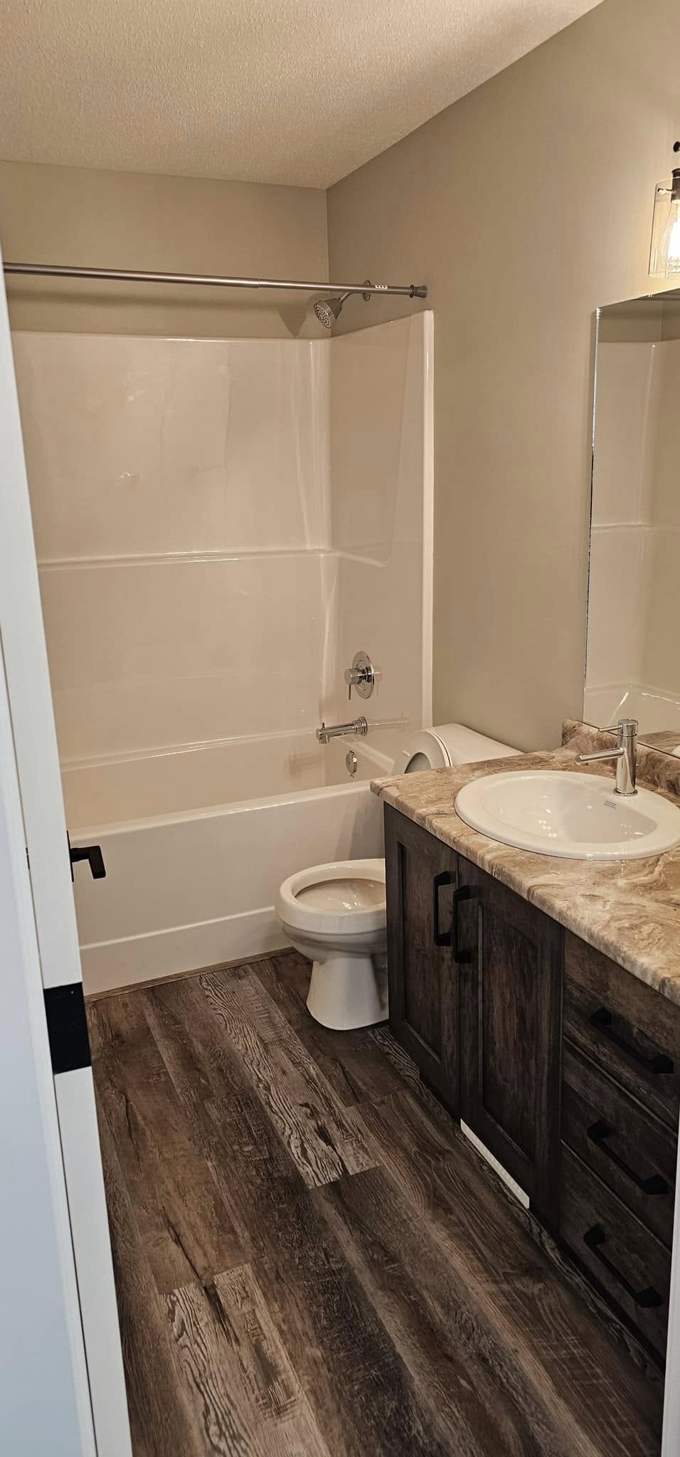 A modern bathroom features a white tub-shower combo, beige walls, dark wood vanity with a marble countertop, sink, and mirror. Rustic wood floors add warmth.