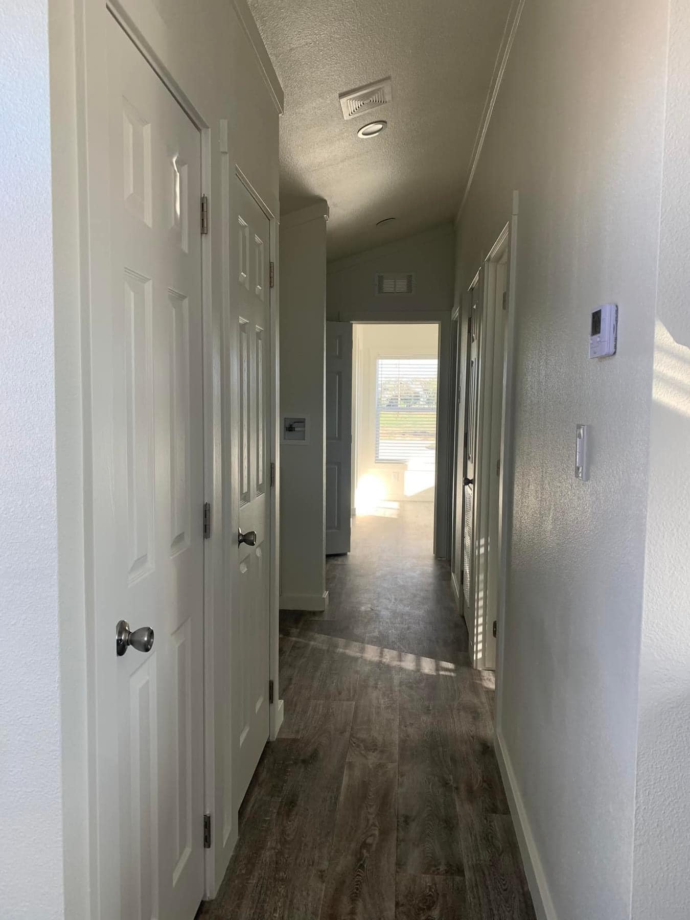 Narrow hallway with white doors on both sides leads to a sunlit room with large windows. Wooden floor adds warmth, evoking a calm, inviting feel.