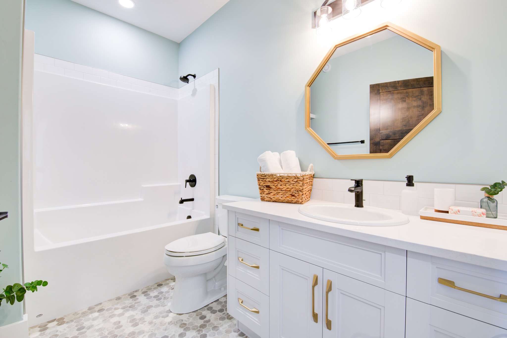 A bright bathroom with blue walls features a white bathtub, toilet, and vanity. A hexagonal mirror and basket with towels add warmth and elegance.