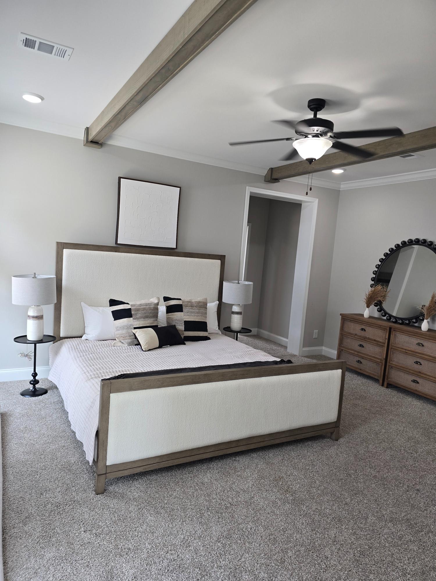 Modern bedroom with a wooden and upholstered bed, neutral bedding, decorative pillows. Ceiling fan, lamps, dresser, mirror, and exposed beams create a cozy, elegant feel.