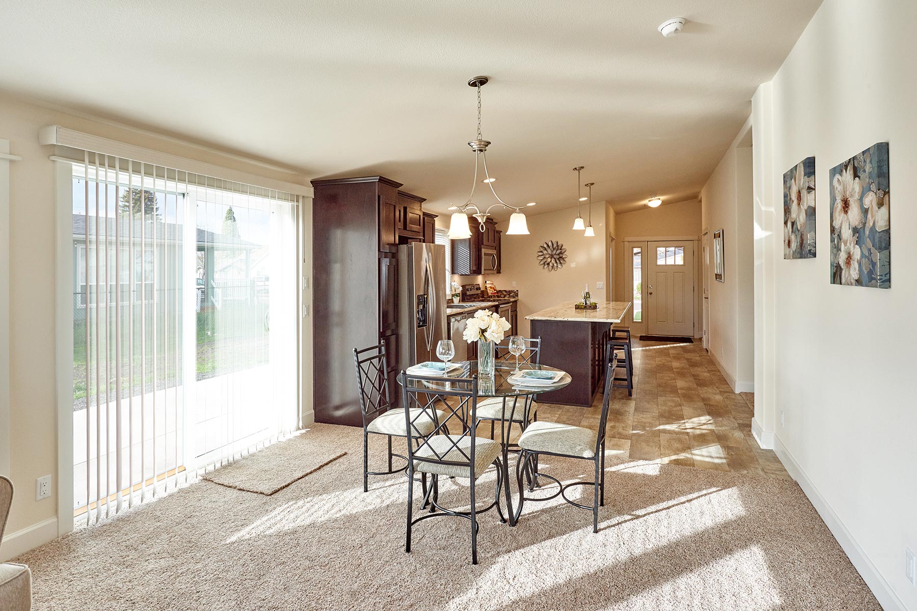 Bright dining and kitchen area with beige carpet, round glass table, and four chairs. Natural light streams through vertical blinds.