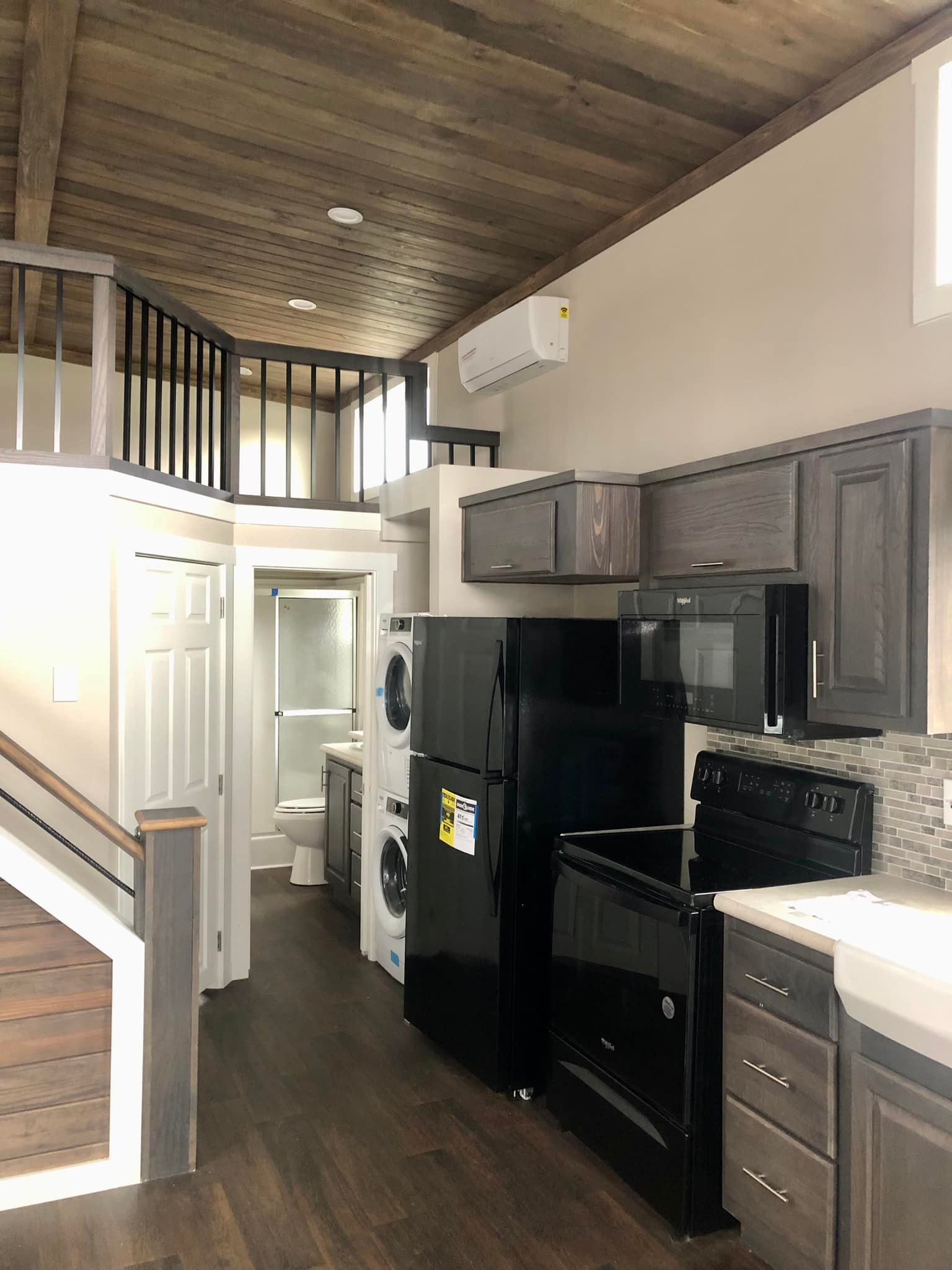 Modern kitchen with dark wood cabinets, black appliances, and wood-paneled ceiling. A small staircase leads to a loft above a laundry and bathroom area. Cozy and compact.