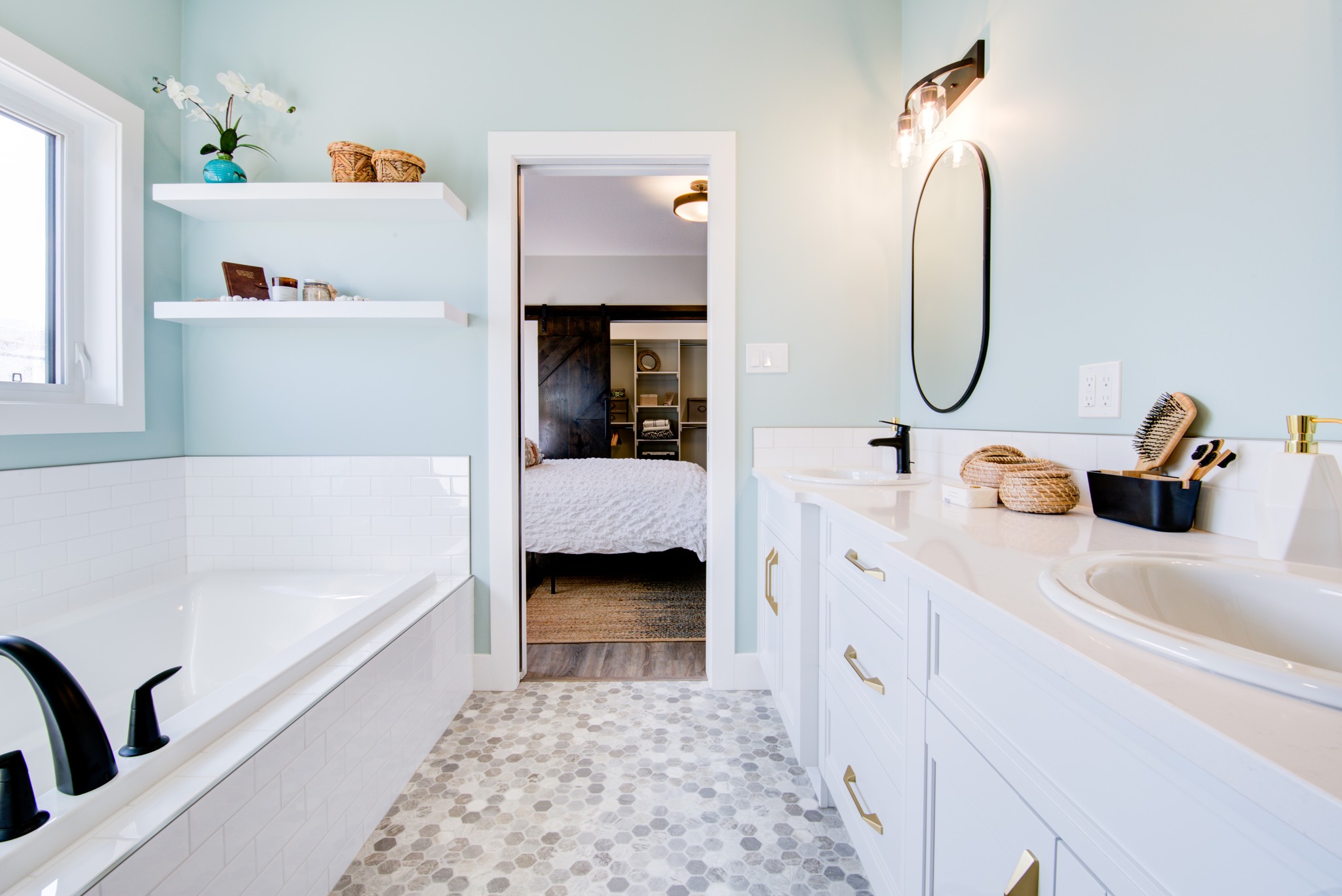 Bright bathroom with light blue walls and patterned floor tiles. Features a bathtub with black fixtures, double sinks, oval mirror, and view into a cozy bedroom.