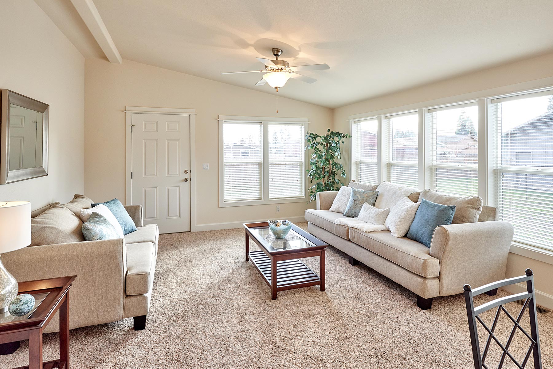 A bright living room with beige sofas, blue cushions, and a glass coffee table on a carpeted floor. Large windows let in natural light, creating a cozy atmosphere.