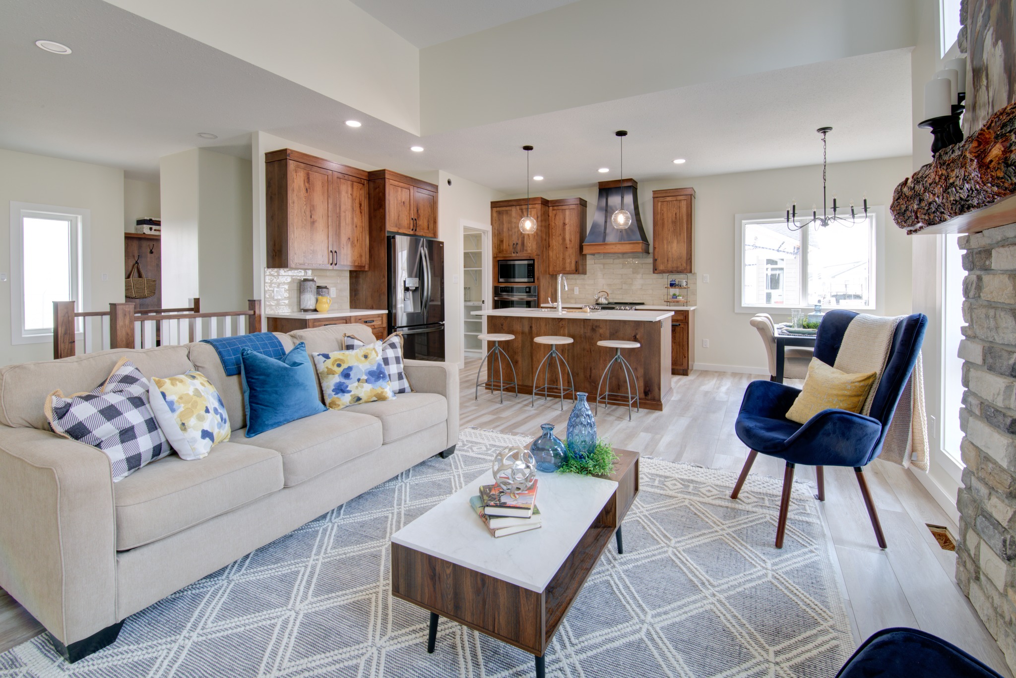 Modern open-concept living room and kitchen. Beige sofa with blue and patterned pillows, wooden cabinets, island with stools, blue armchair, and stylish decor.