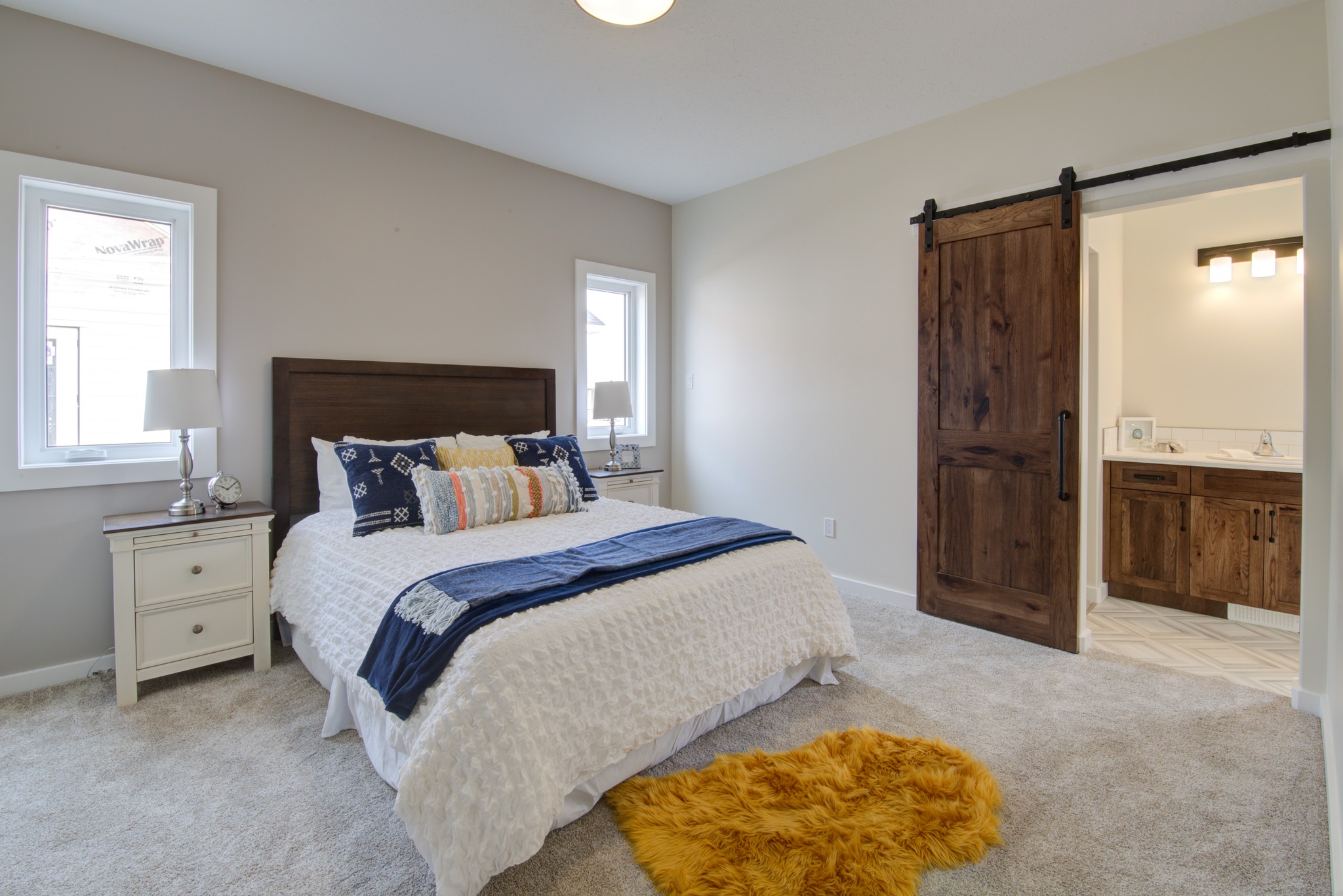 A cozy bedroom with a neatly made bed featuring blue and white pillows. Two lamps adorn white bedside tables. A wooden barn door leads to a bathroom. A yellow rug adds a pop of color on the gray carpet, creating a warm and inviting atmosphere.
