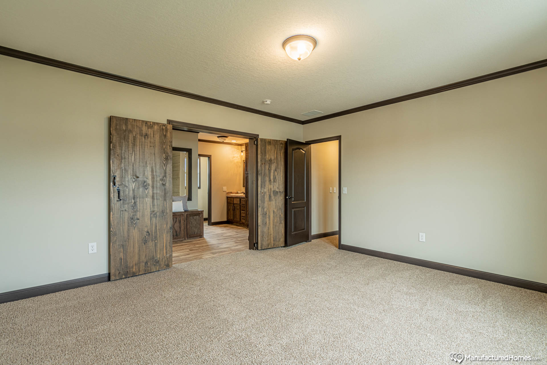 Spacious beige bedroom with carpet, rustic wood double door opening to a lit bathroom. Simple, warm, and inviting ambiance with dark wood trim.