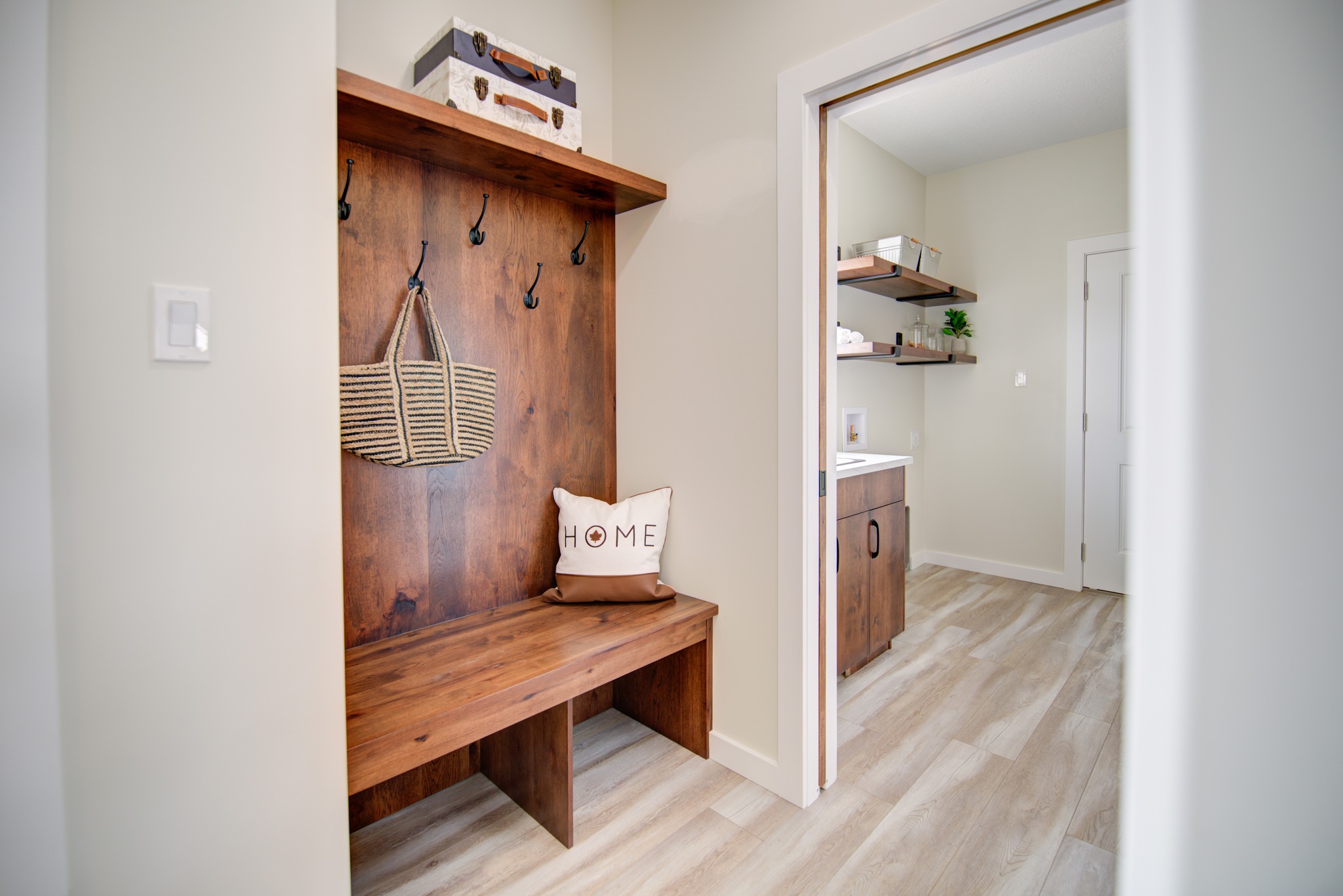 A cozy mudroom features a wooden bench with hooks above, holding a woven bag. A "HOME" cushion sits on the bench. Adjacent is a clean laundry room.
