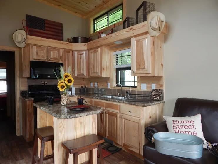 Rustic kitchen with natural wood cabinets, a small island with stools, and a sunflower centerpiece. Cozy living area with a "Home Sweet Home" pillow.