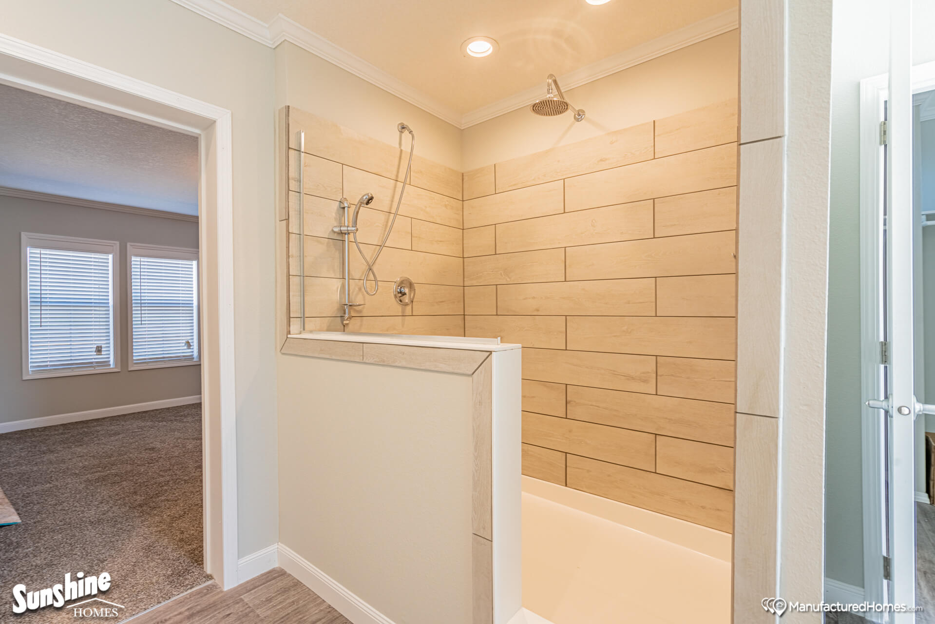 Spacious bathroom with a large beige-tiled shower featuring a rainfall showerhead. Adjacent room has carpet flooring and two bright windows. Calm, modern feel.