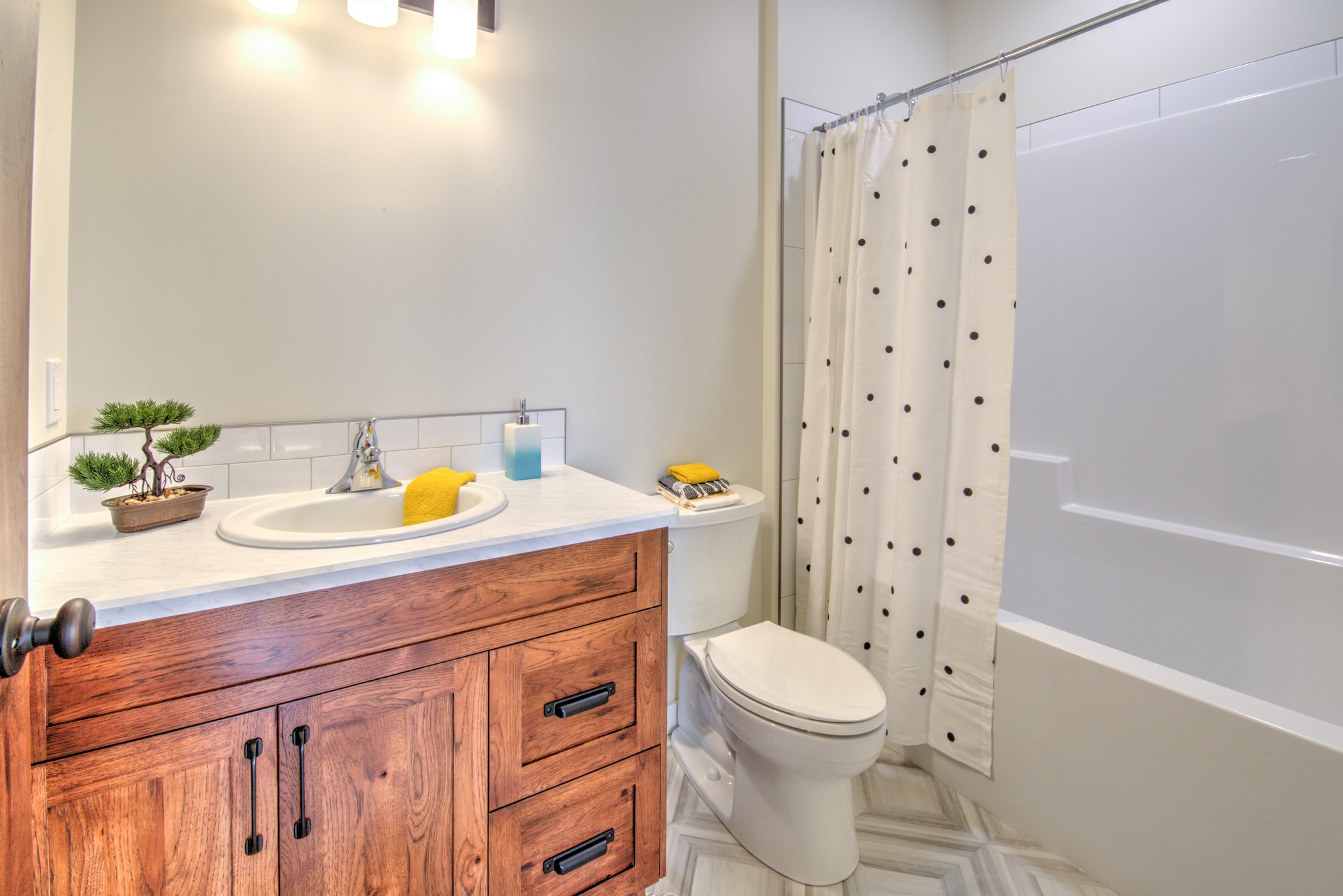A cozy bathroom with a wooden vanity and white countertop. A bonsai plant and soap sit beside the sink. A bathtub features a polka dot shower curtain.