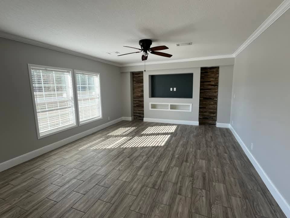A spacious, empty room with light gray walls, white baseboards, and wood flooring. Large windows let in sunlight, highlighting a ceiling fan.