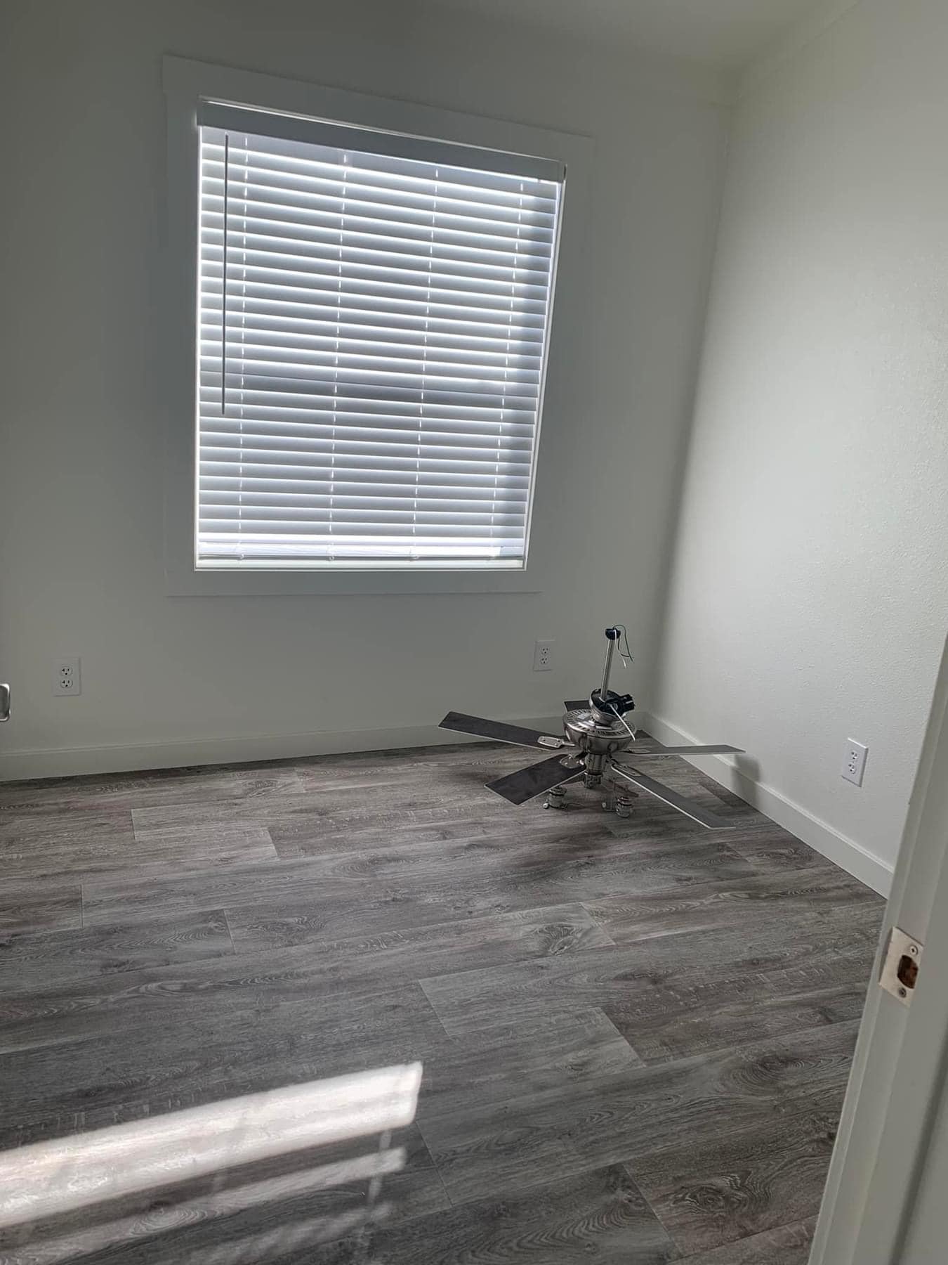 Empty room with white walls and a window with closed blinds on the left. A detached ceiling fan rests on the wood-look floor, creating an unfinished, bare atmosphere.