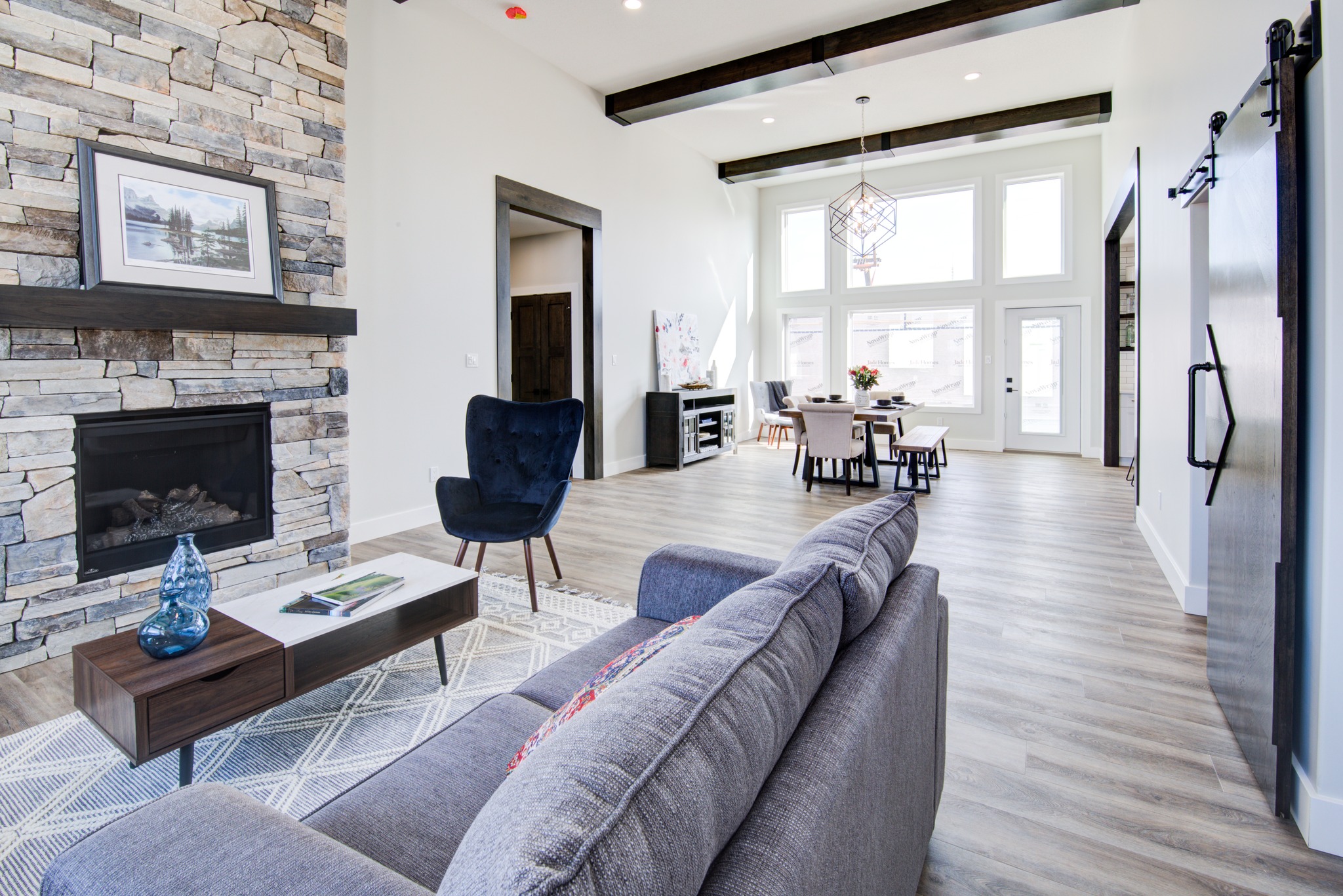 Modern living room with light wood floors, gray sectional, and stone fireplace. Large windows and dining area create a bright, open atmosphere. Calm and inviting.