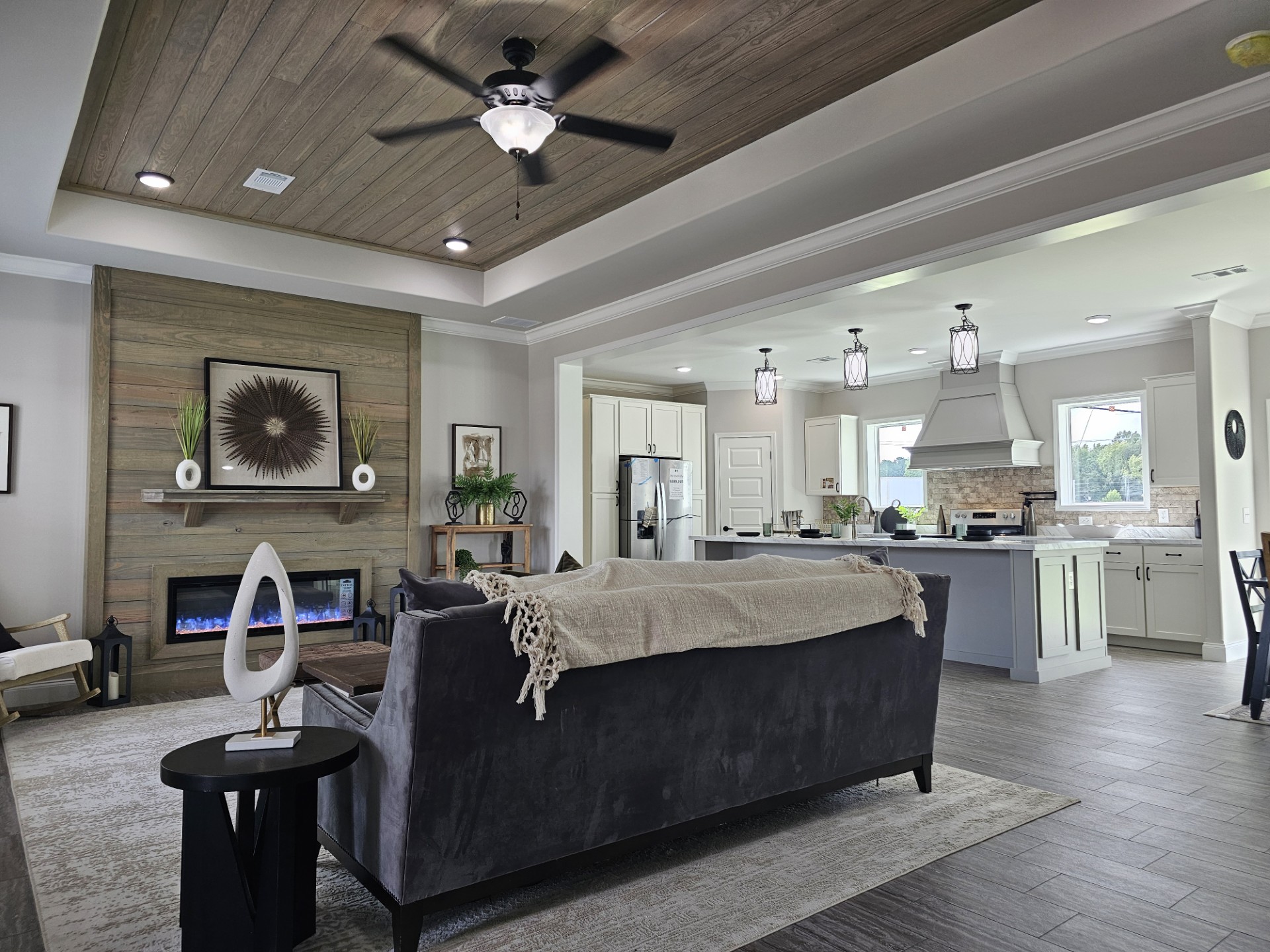 Spacious living room with a wooden ceiling and fan, gray sofa, and modern fireplace. Open kitchen with pendant lights. Calm, inviting atmosphere.