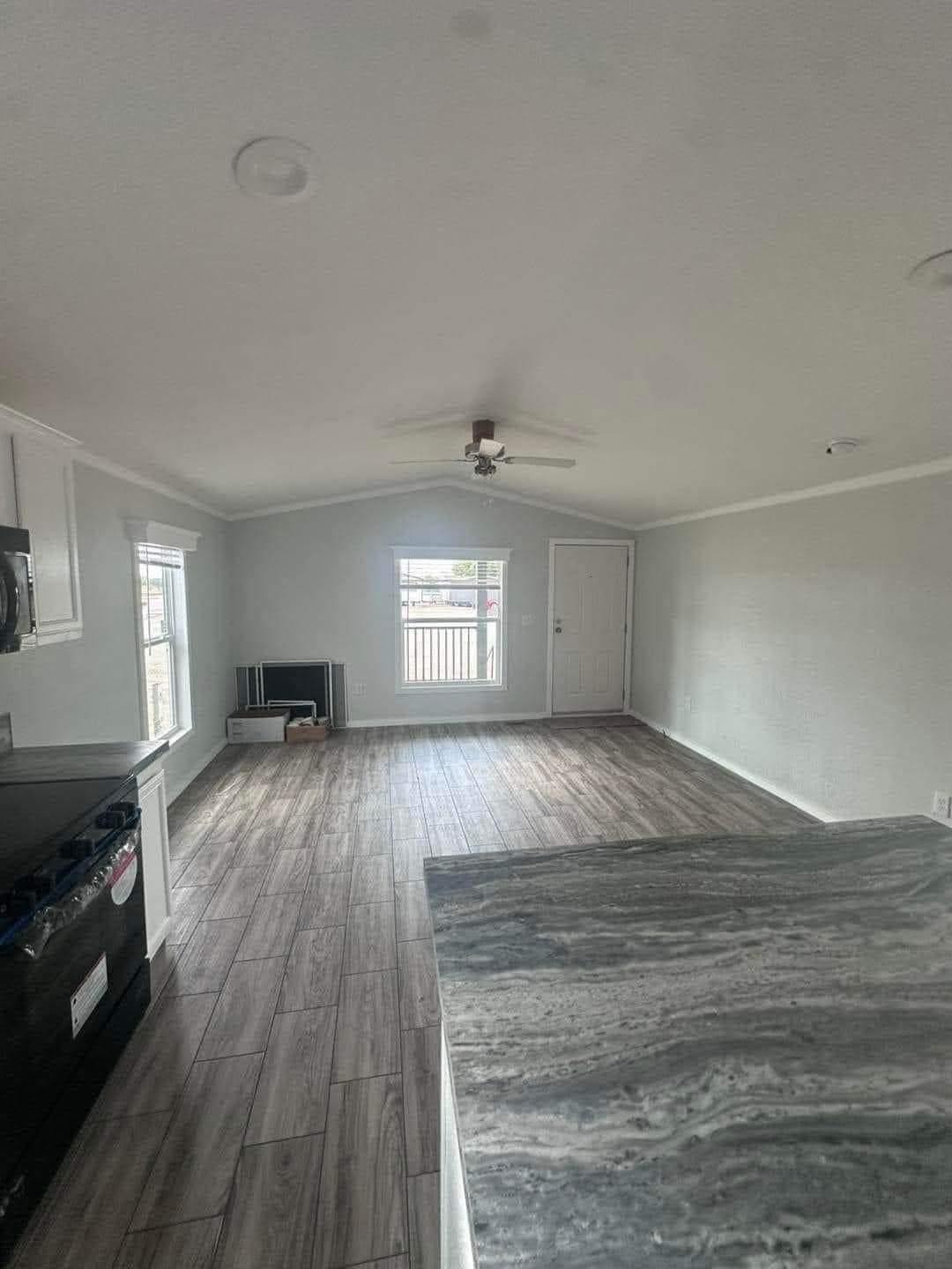 A bright, empty room with light gray walls and wood flooring. A window and door are at the far end, under a ceiling fan. Kitchen area is visible on the left.