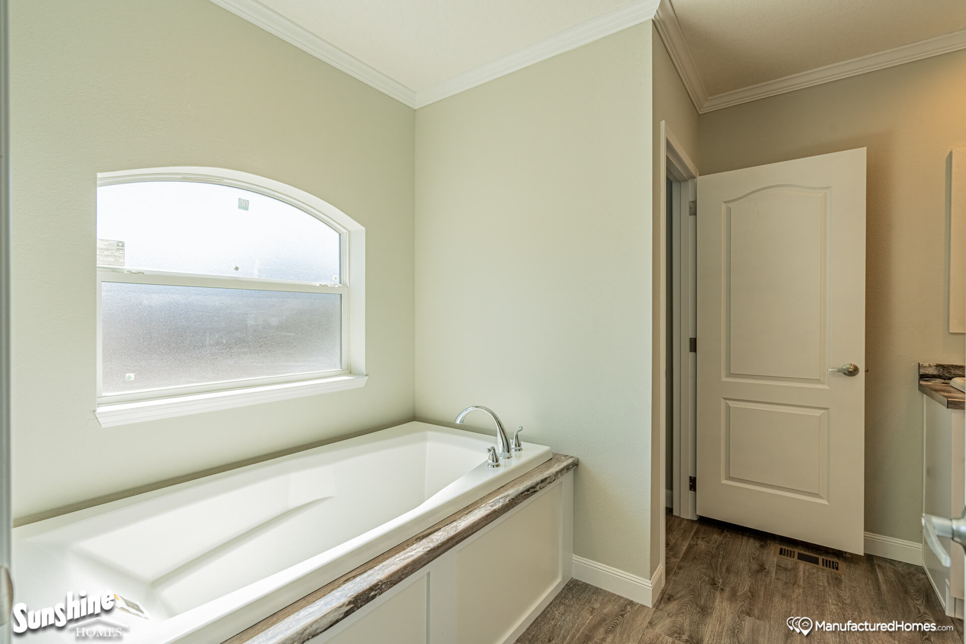 Bright bathroom with a large white bathtub under an arched window, light beige walls, and wood-textured flooring, creating a serene atmosphere.