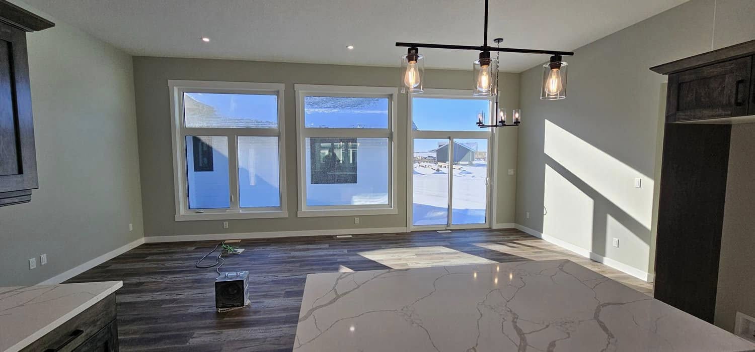 Spacious, sunlit living area with large windows and glass doors overlooking snowy landscape. Modern chandelier hangs above polished wood floor, creating a welcoming ambiance.