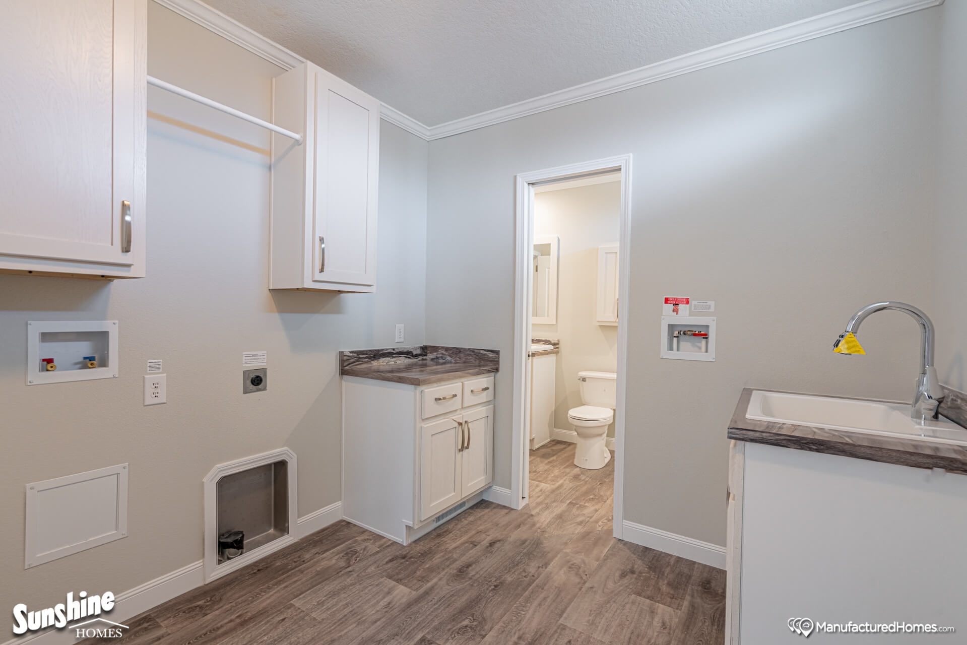 Laundry room with light gray walls, wood flooring, white cabinets, and a countertop. A door on the left leads to a white bathroom.