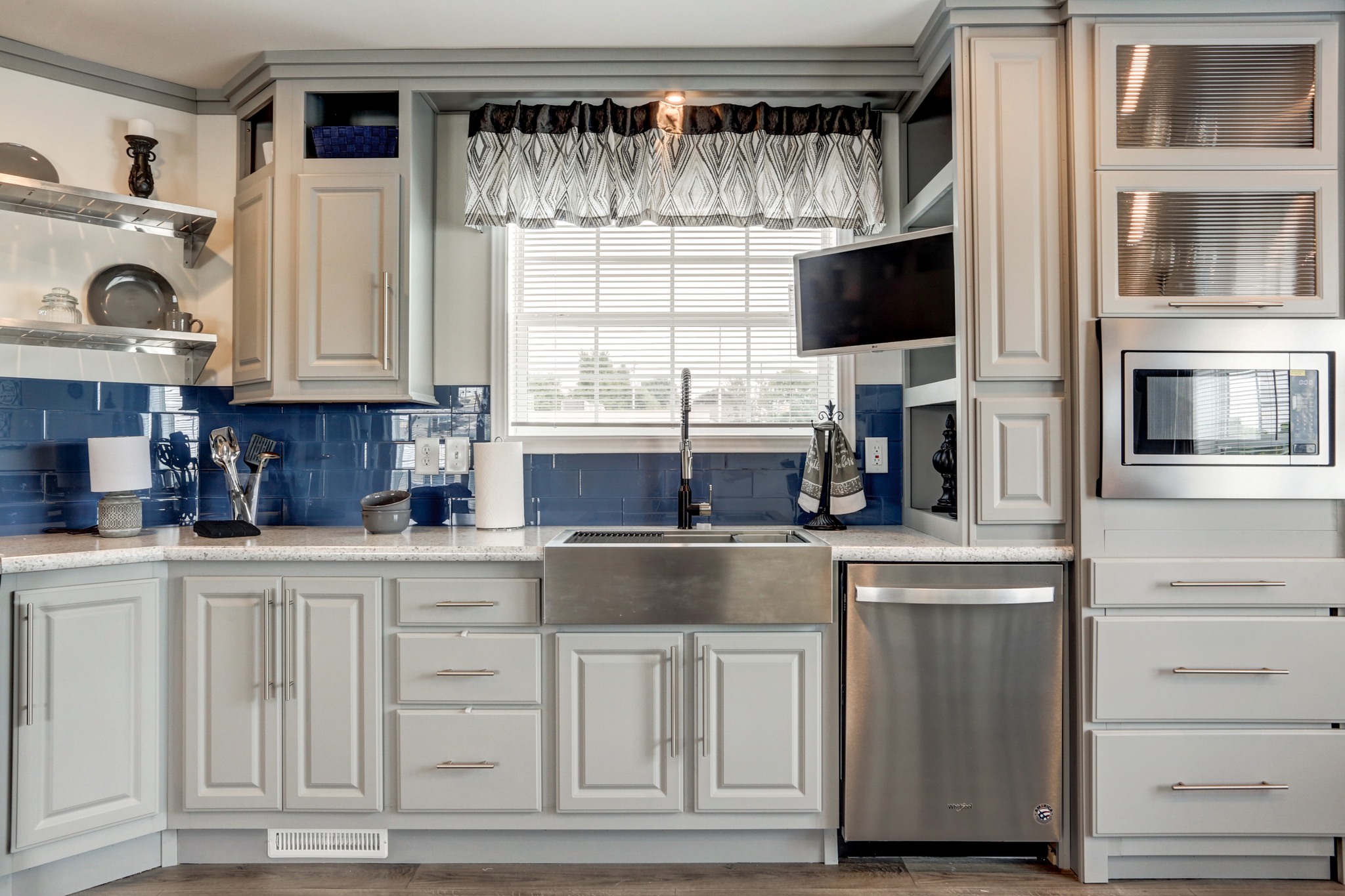 Bright kitchen with modern gray cabinets and stainless steel appliances. A blue tile backsplash contrasts with white countertops. A window with patterned curtains adds a cozy touch.