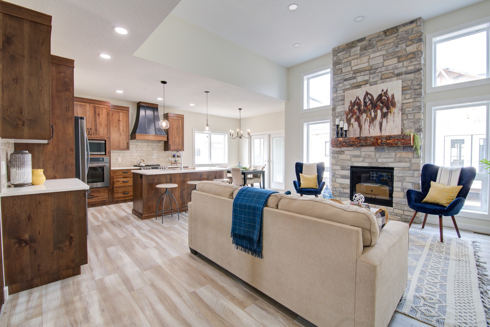 Spacious modern living room with a stone fireplace, cozy beige sofa, blue chairs, and a kitchen with wooden cabinets. Bright and inviting atmosphere.