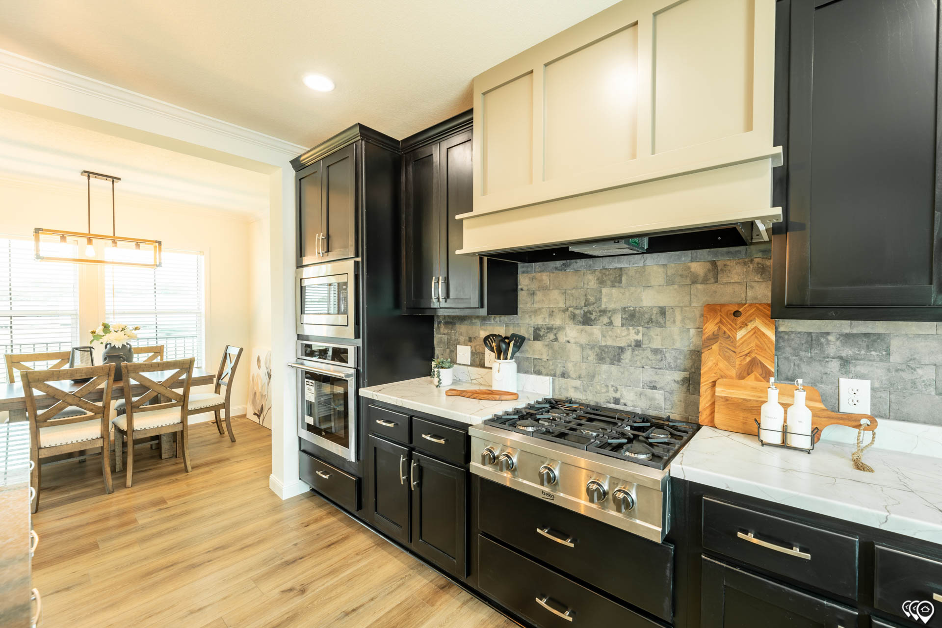Modern kitchen with black cabinets, stainless steel appliances, and stone backsplash. A dining area with wooden table and chairs is visible nearby.