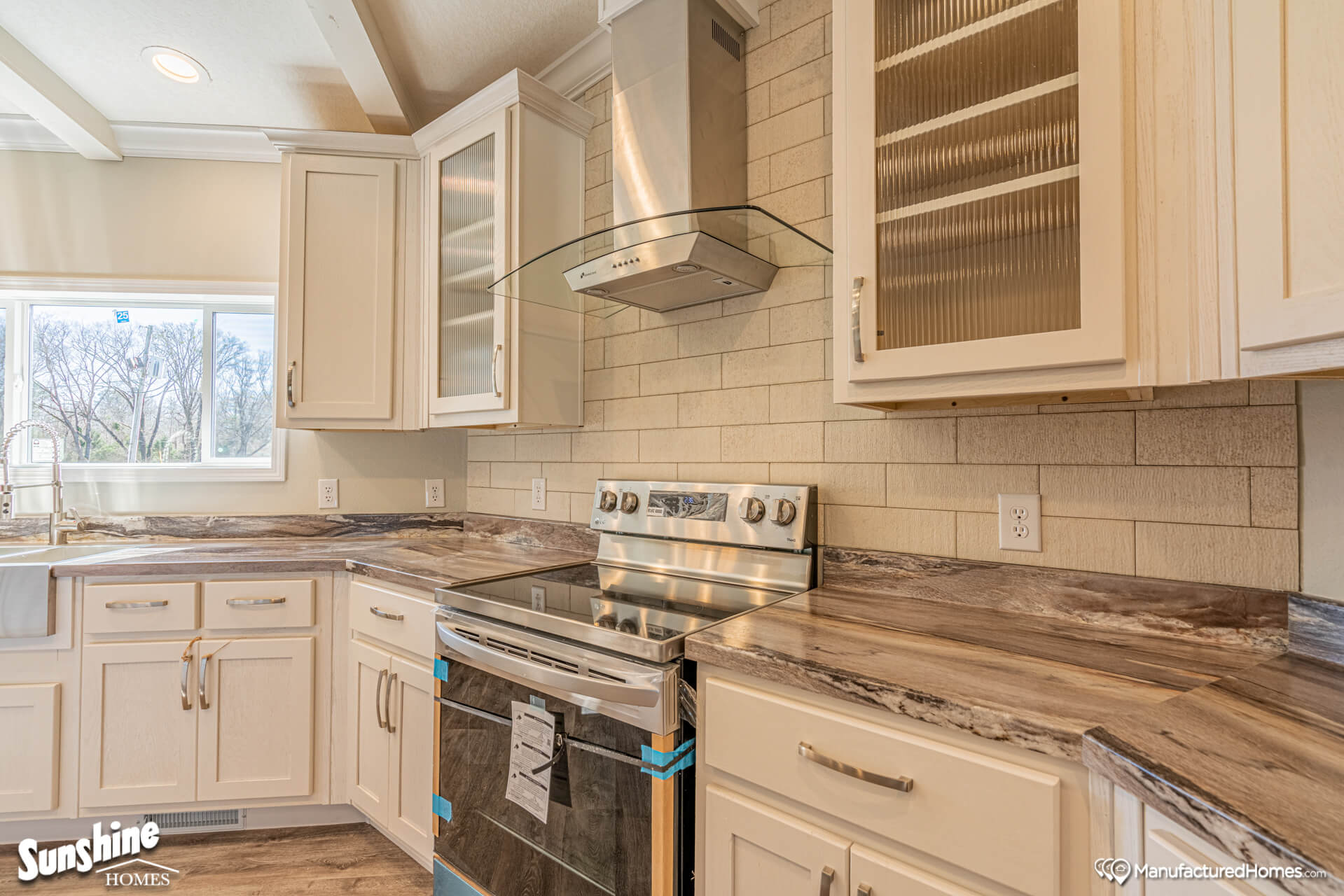 Bright, modern kitchen with white cabinets, stainless steel range, glass hood, and marbled countertops. Sunlight streams through a large window.