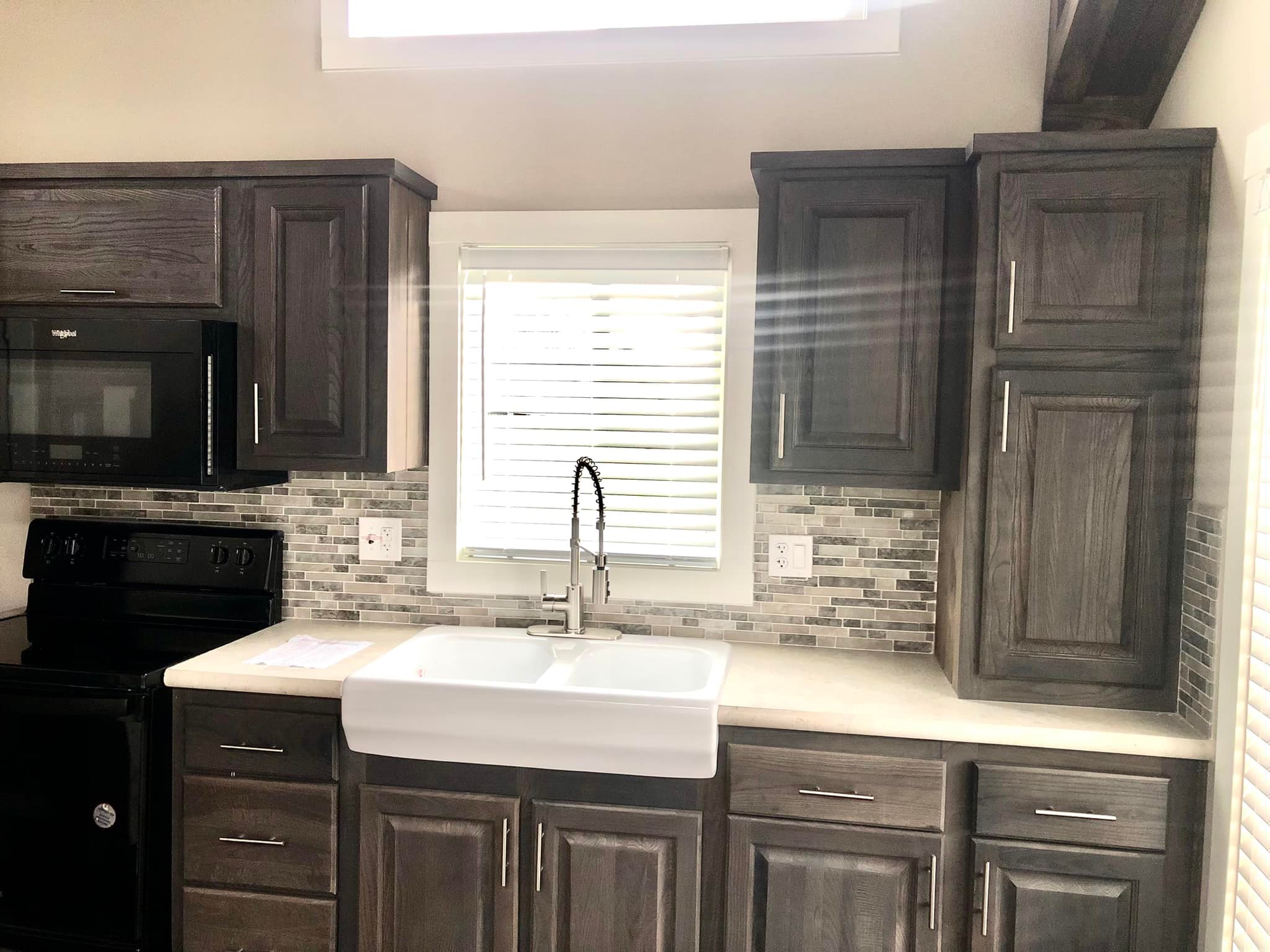 Modern kitchen with dark wood cabinets and light countertops. Featuring a farmhouse sink under a window with blinds, mosaic tile backsplash, and black appliances. Bright, clean, and inviting atmosphere.