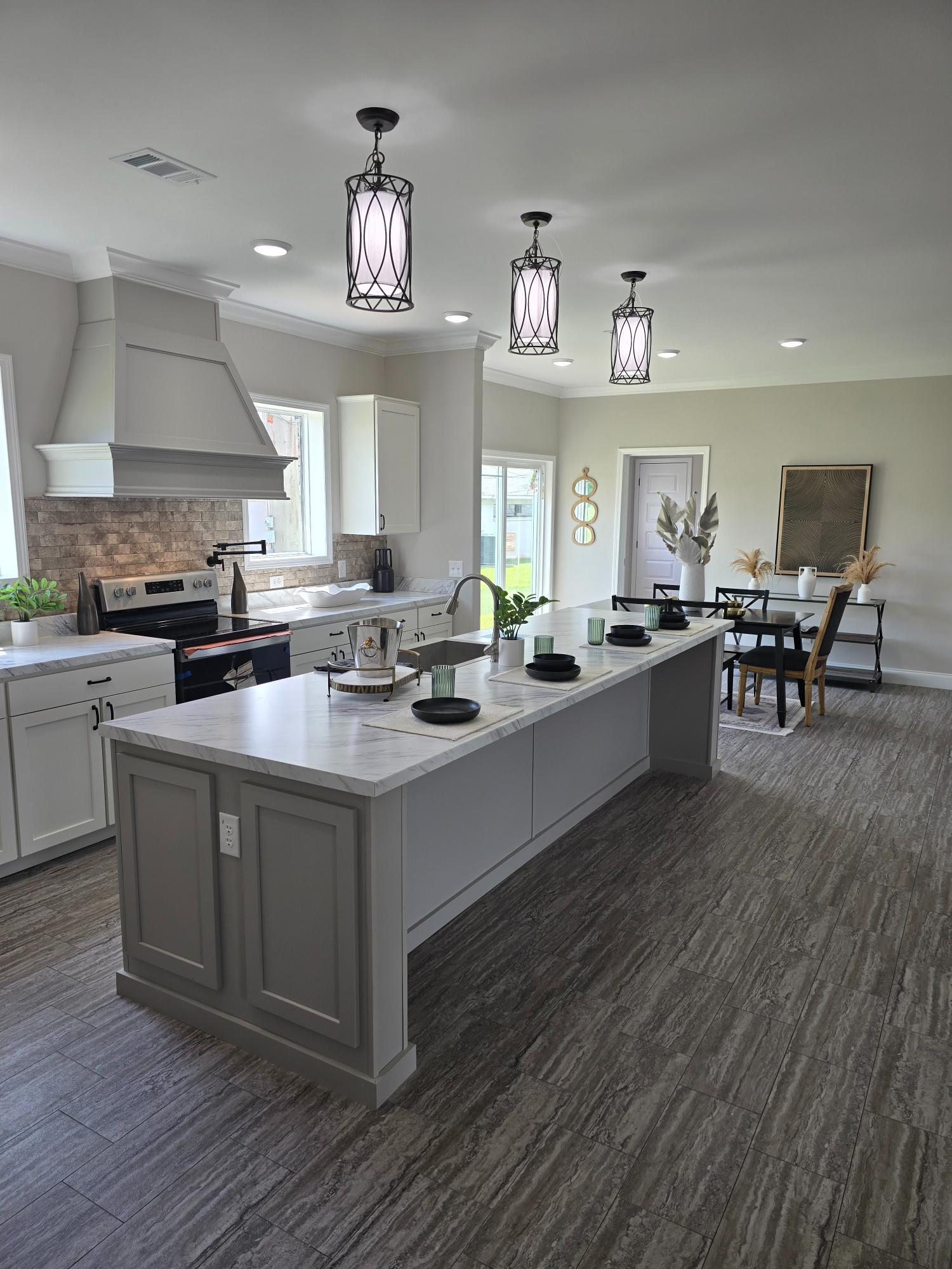 Spacious kitchen with a gray island, white cabinets, and pendant lights. Dining area in the background features a table with decorative plants.