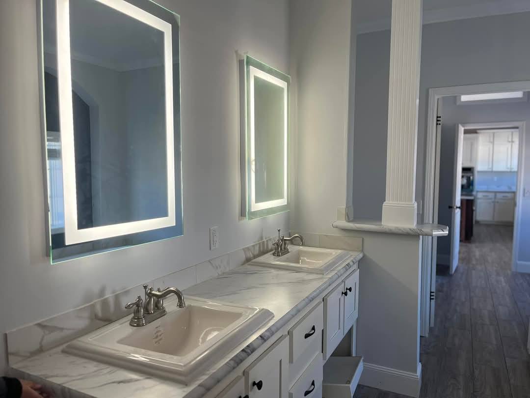 Modern bathroom with two illuminated mirrors above a marble countertop with dual sinks. White cabinetry and wood floors add a clean, elegant feel.