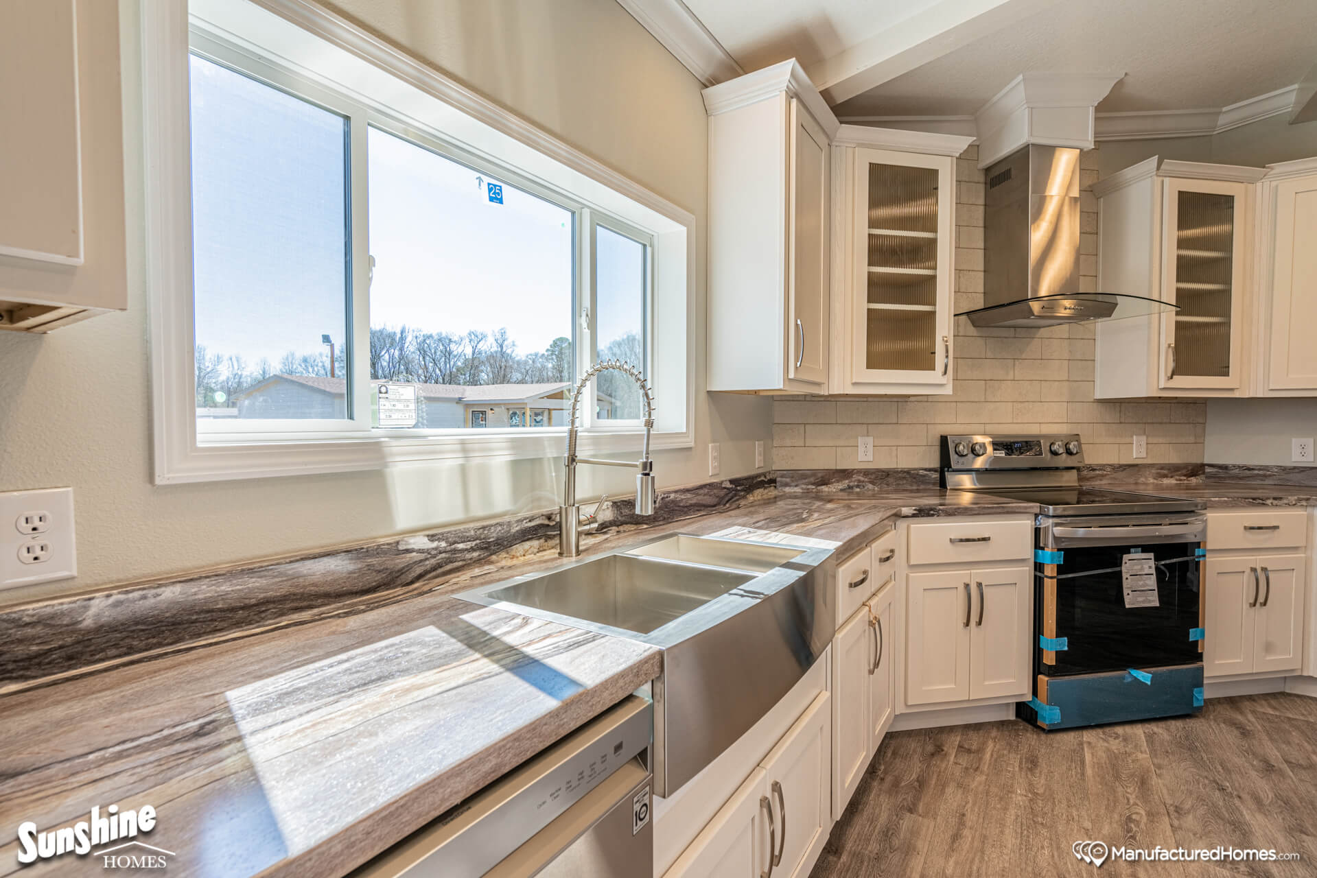 Modern kitchen with a sleek stainless steel sink, marbled countertops, white cabinets, and a stainless steel stove. Bright light from a large window.