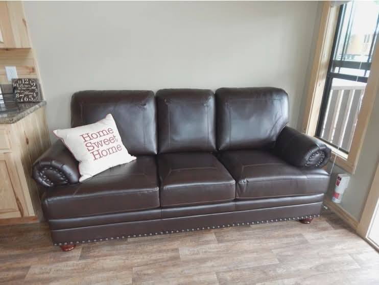A dark brown leather sofa with a "Home Sweet Home" pillow sits against a beige wall. The room has wooden flooring and a large window on the right.