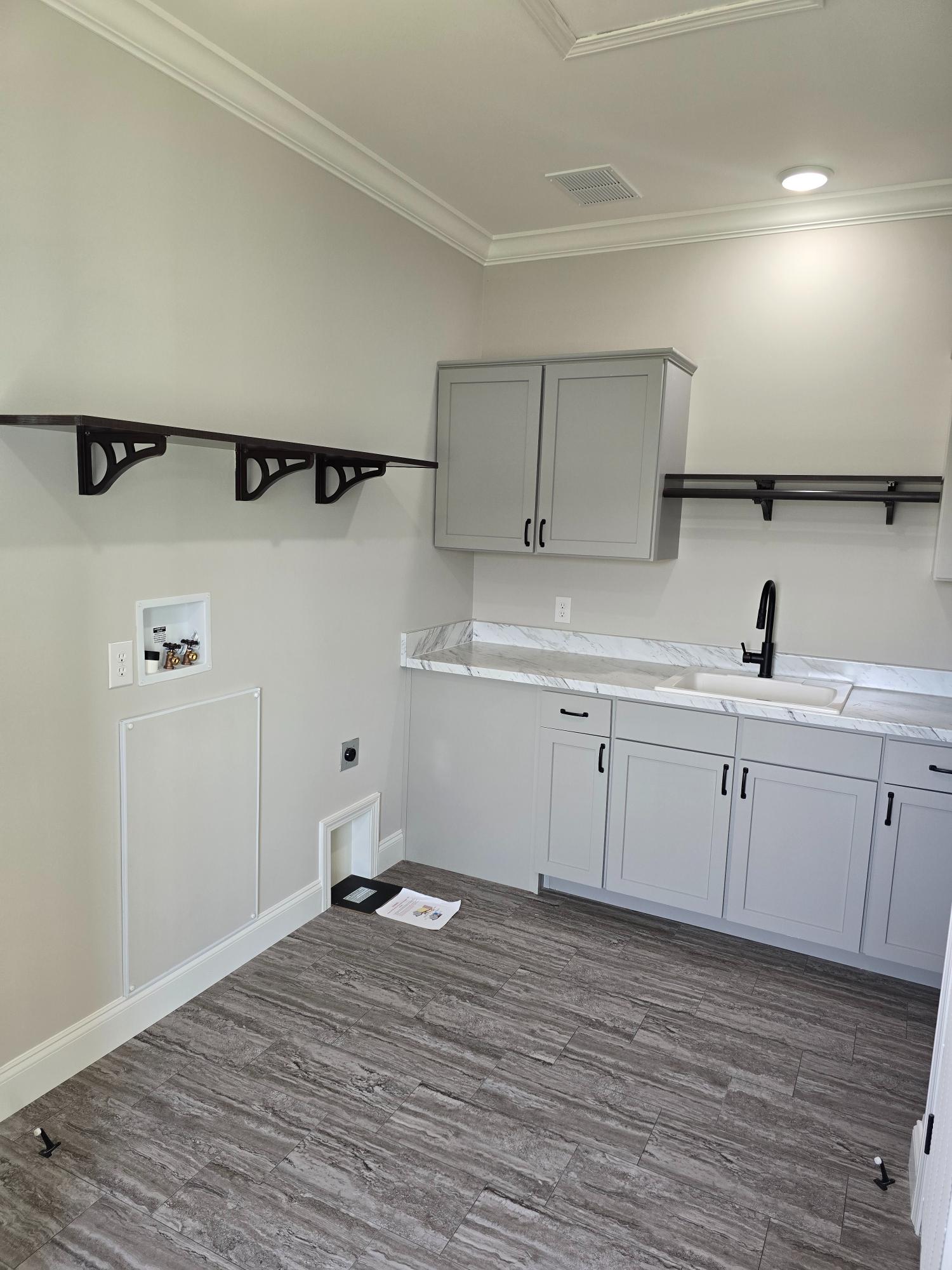 A modern laundry room with gray cabinets and a marble countertop. Black faucet and handle accents, open shelving, and wood-patterned tile flooring.