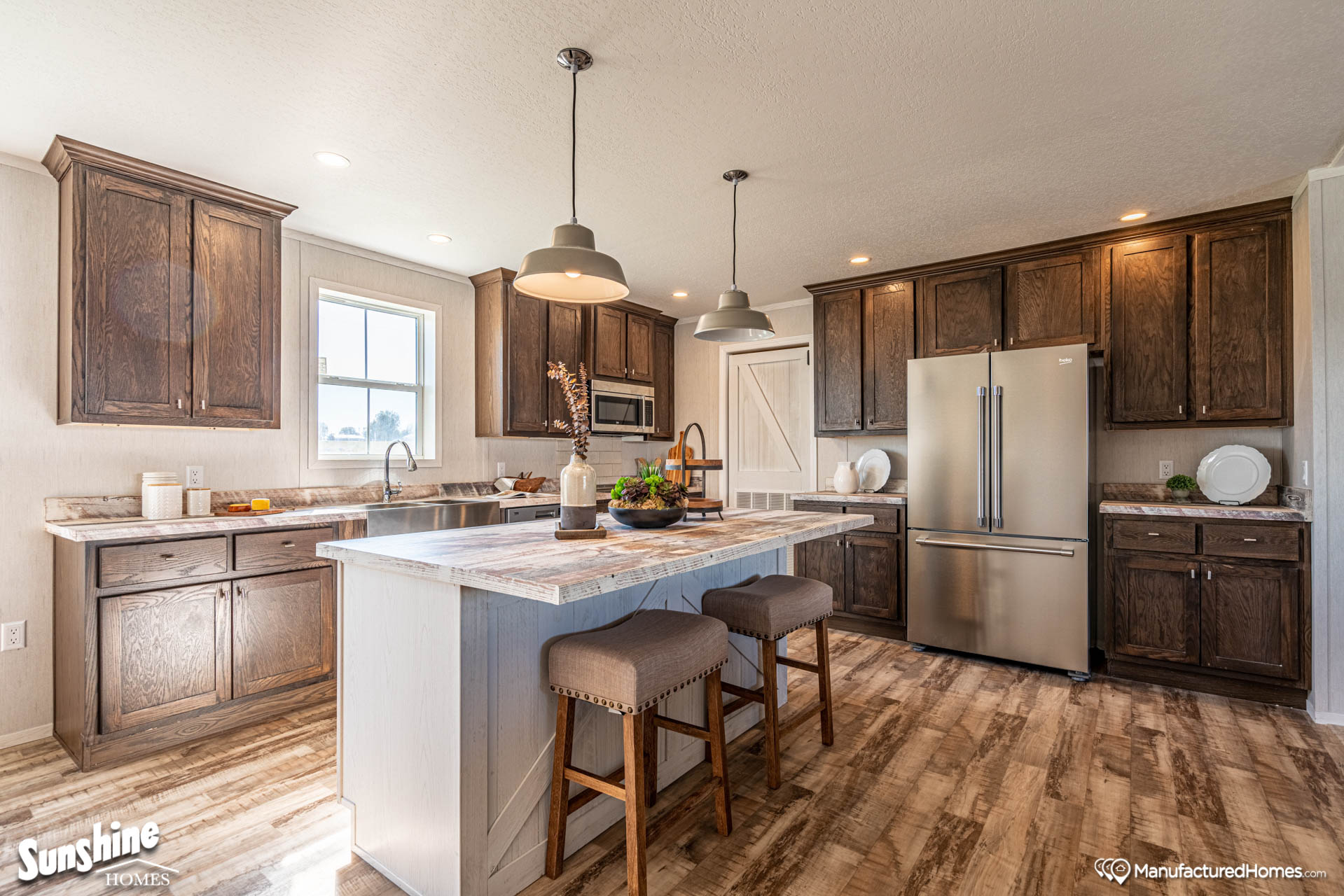 Spacious kitchen with wooden cabinets, a large island with stools, and a stainless steel fridge. Warm lighting creates a cozy, inviting atmosphere.