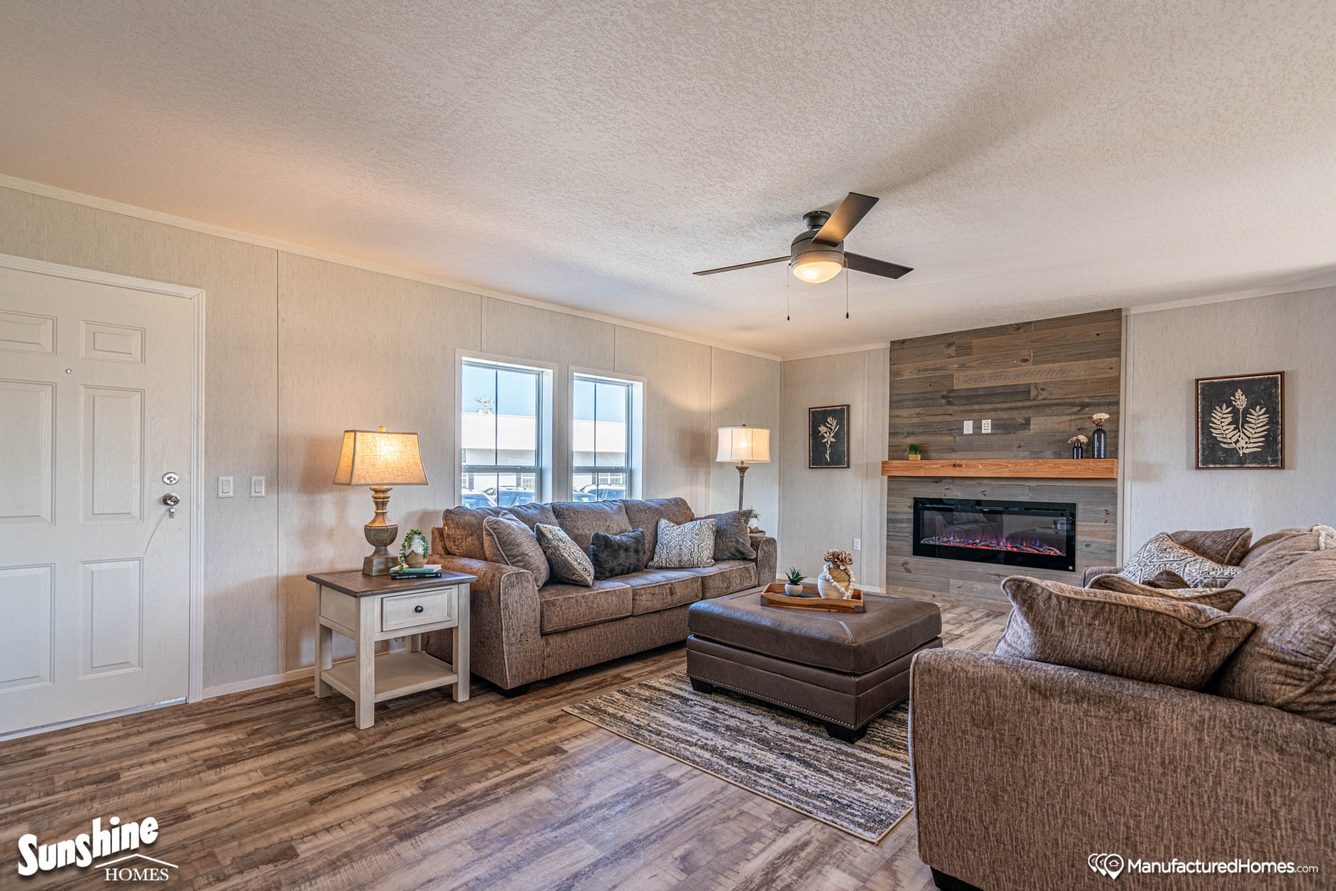 Cozy living room with two beige sofas, a dark ottoman, and a wooden accent wall featuring an electric fireplace. Warm lighting enhances the inviting atmosphere.