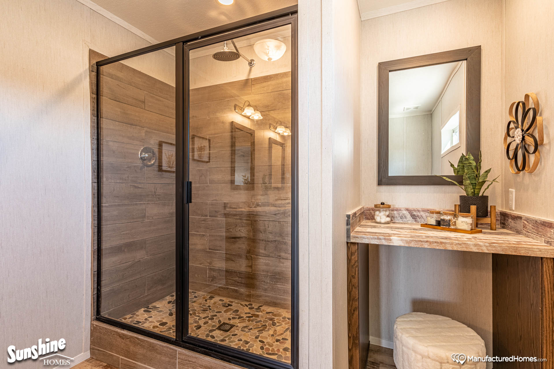 Modern bathroom with a glass-enclosed shower featuring stone tiles and a rainfall showerhead. Adjacent is a wooden vanity with a mirror, plant, and decor.